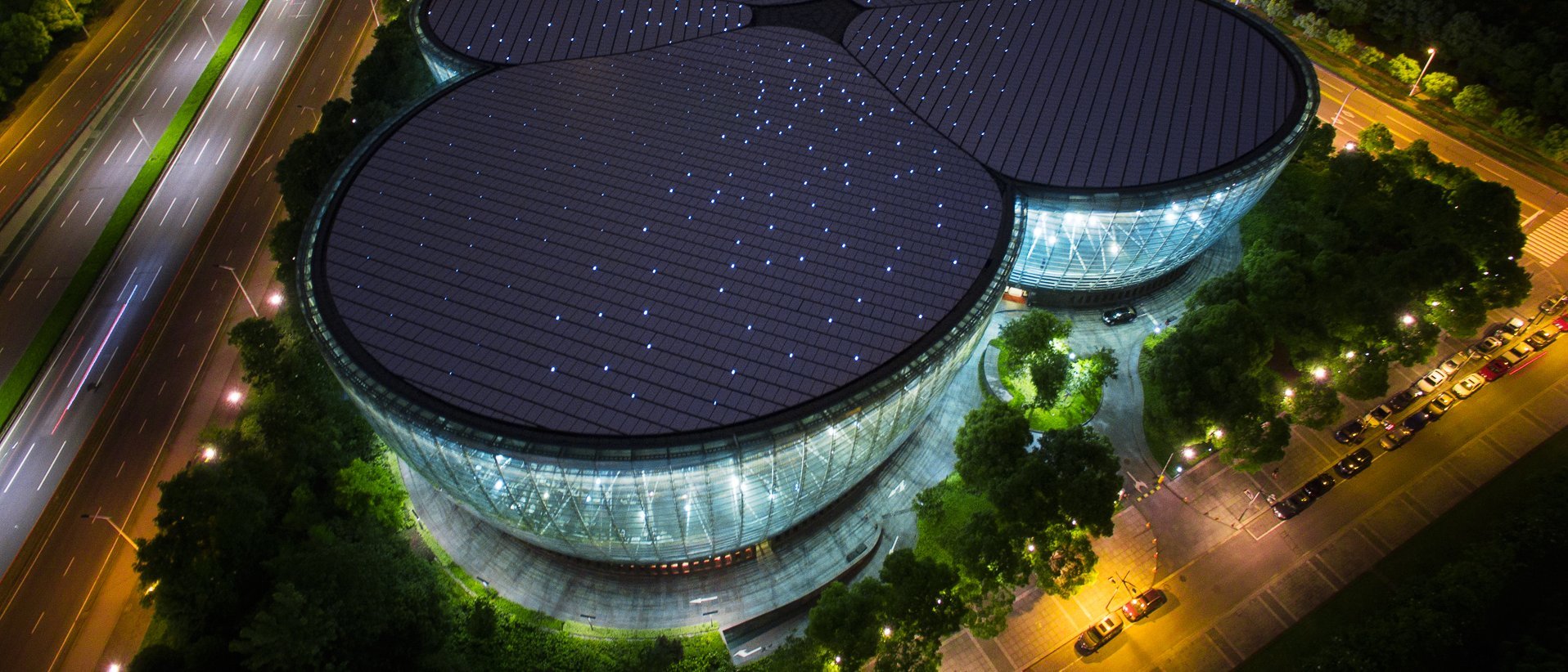 Luftaufnahme eines modernen, einzigartig geformten Gebäudes mit weißem Dach, umgeben von Bäumen und Straßen, in einem städtischen Gebiet mit anderen Gebäuden und Grünflächen in der Nähe.