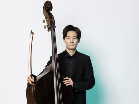 A man dressed in a black suit stands holding a double bass upright with one hand on the instrument’s neck and the other holding a bow, against a white background.