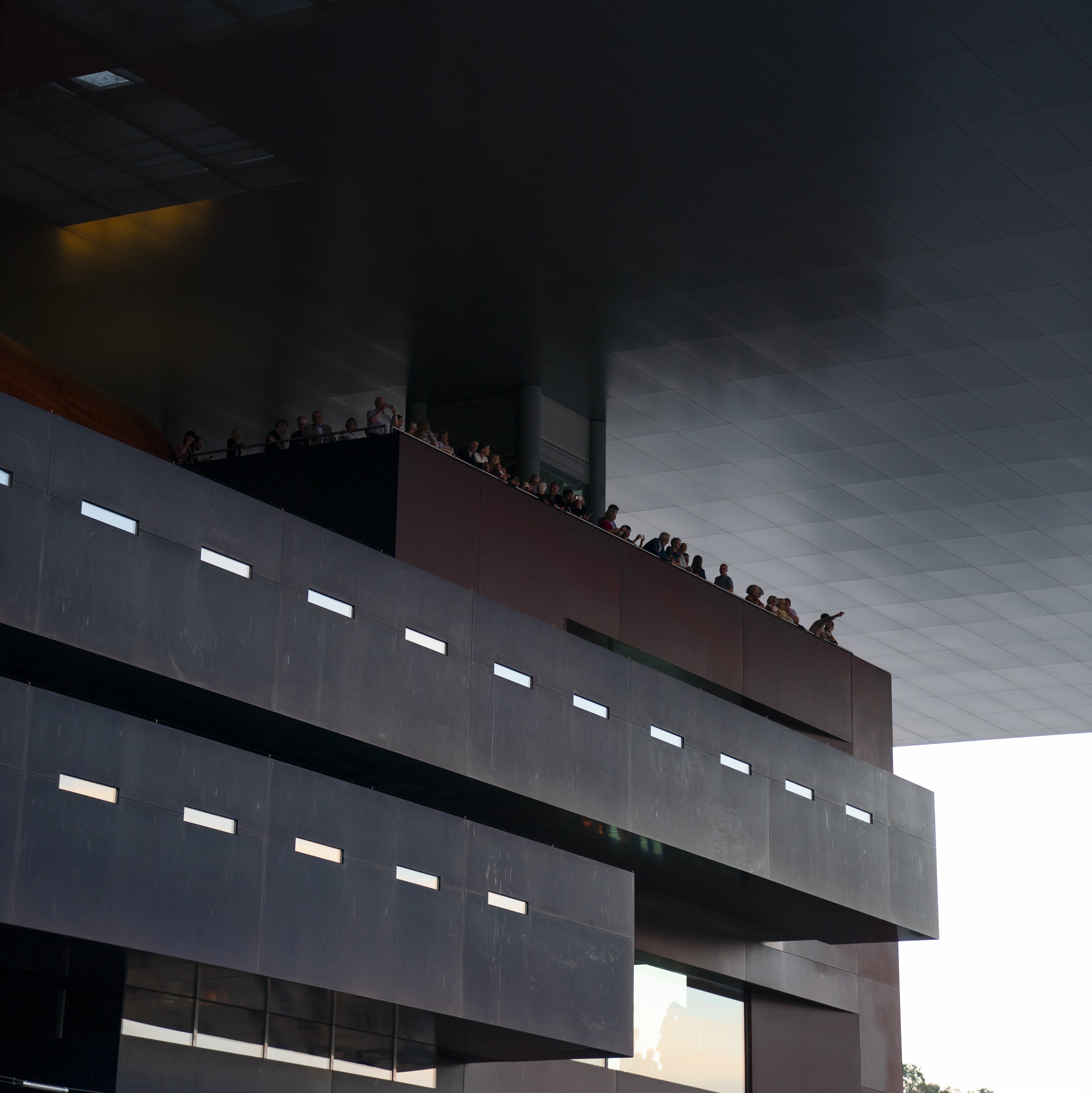People stand on a balcony beneath a large, modern overhanging roof. The dramatic architecture creates sharp lines and deep shadows in the scene.