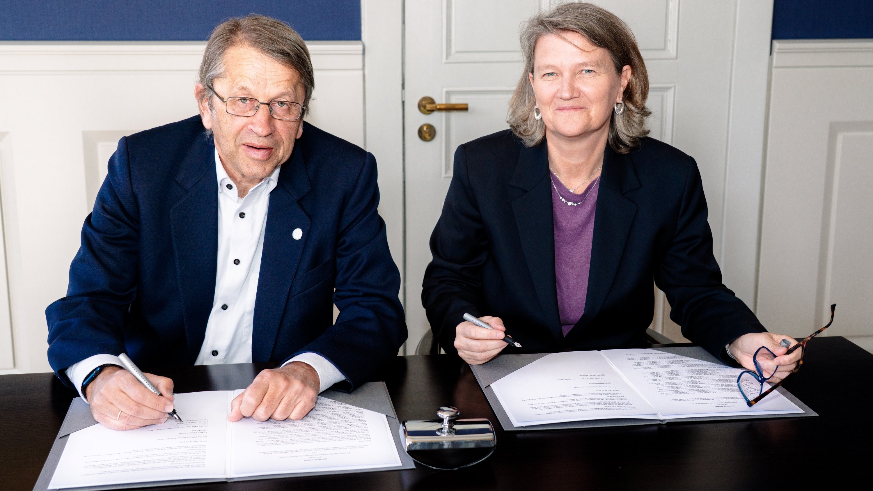 Prof. Heyo K. Kroemer und Andrea Zietzschmann in Geschäftskleidung sitzen an einem Tisch und unterschreiben jeweils ein Dokument. Beide lächeln und schauen in die Kamera. Auf dem Tisch vor ihnen liegen Papiere, ein Stiftständer und eine Brille.