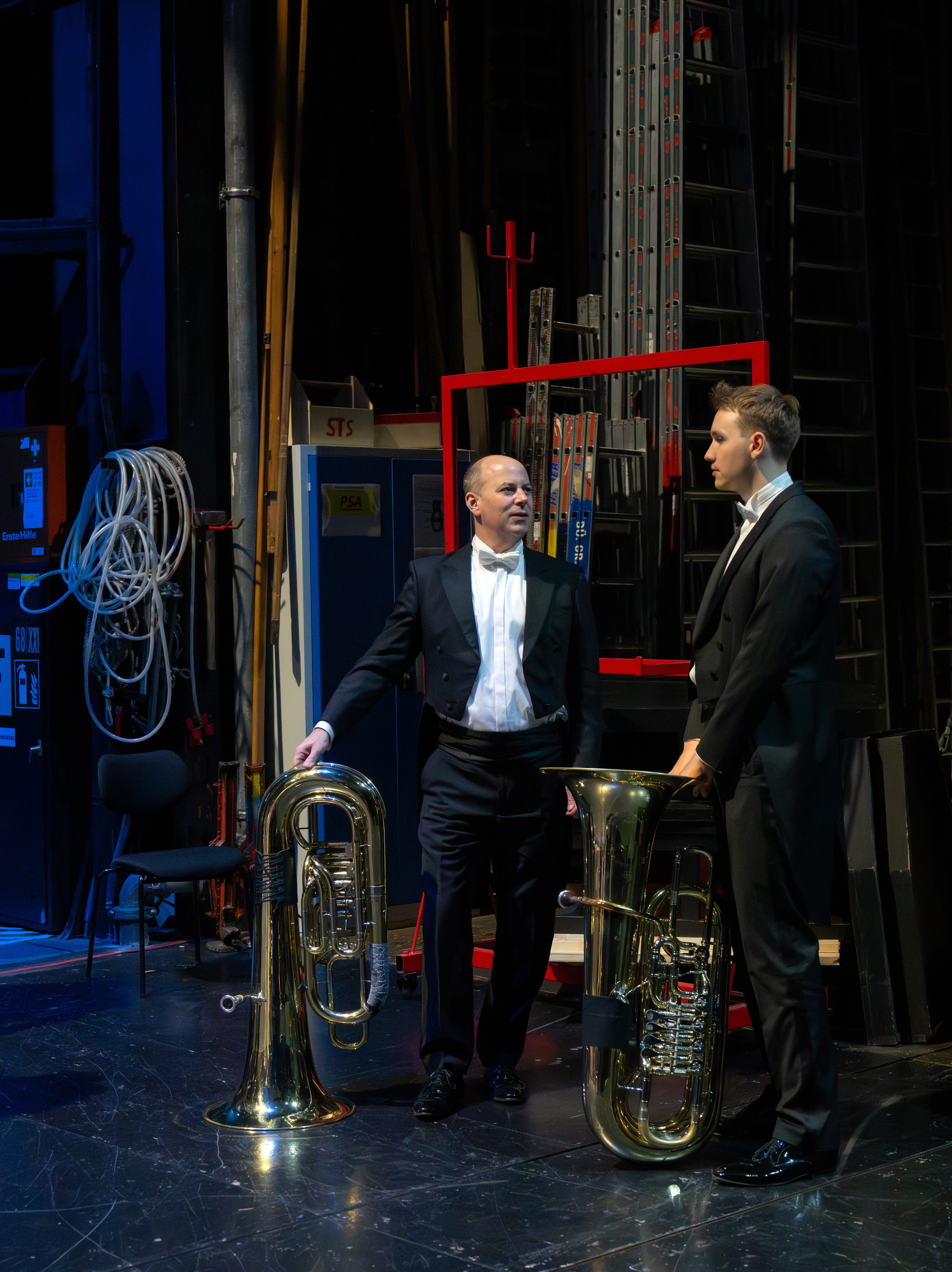 Zwei Männer im Frack stehen hinter der Bühne neben einer Tuba und unterhalten sich. Im Hintergrund sind Kabel, Leitern und Bühnentechnik zu sehen, die eine typische Backstage-Atmosphäre eines Theaters schaffen.