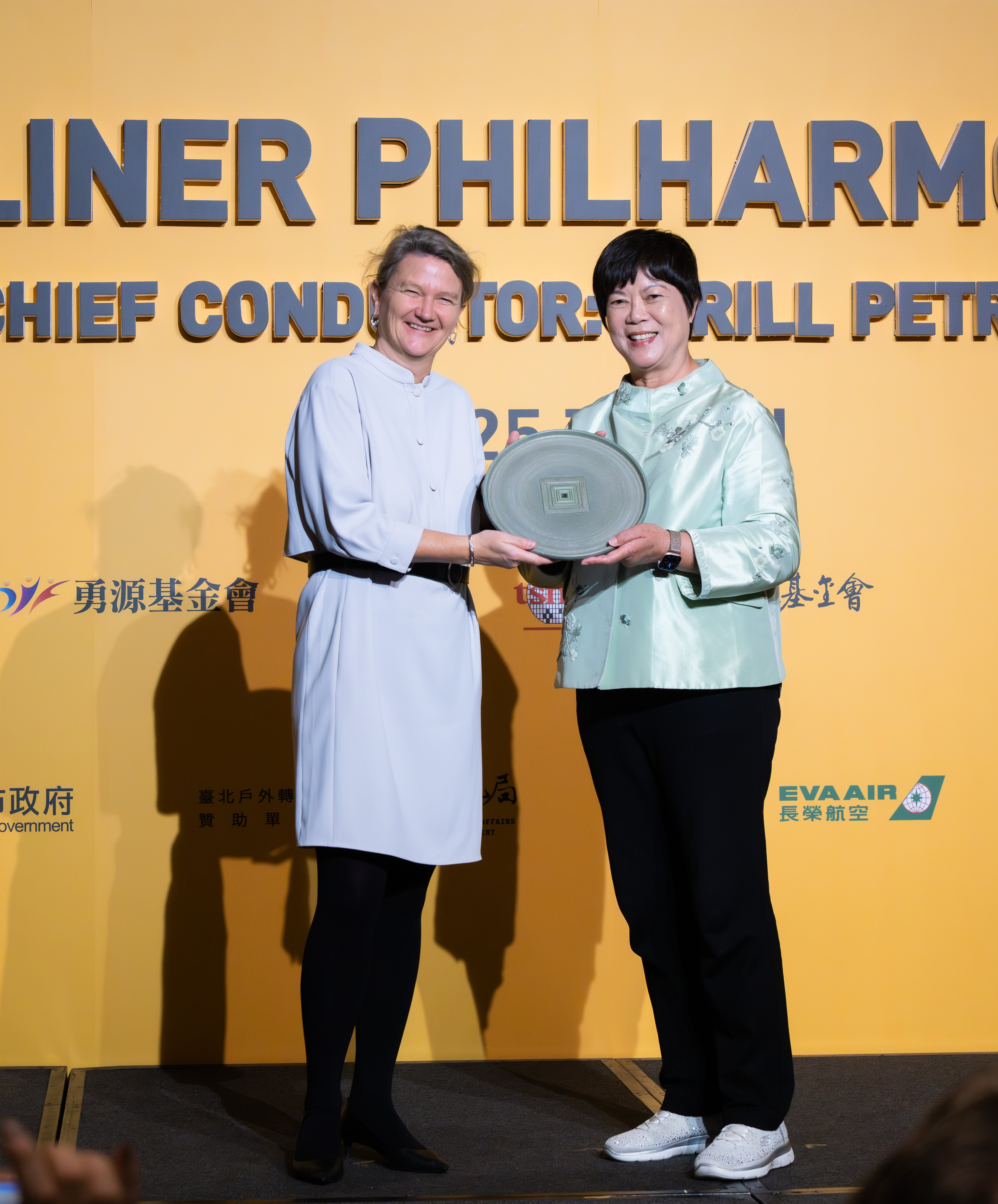 Two women stand smiling on stage, holding a circular glass award together in front of a yellow backdrop with text and logos, dressed in formal attire.