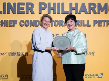 Two women stand smiling on stage, holding a circular glass award together in front of a yellow backdrop with text and logos, dressed in formal attire.
