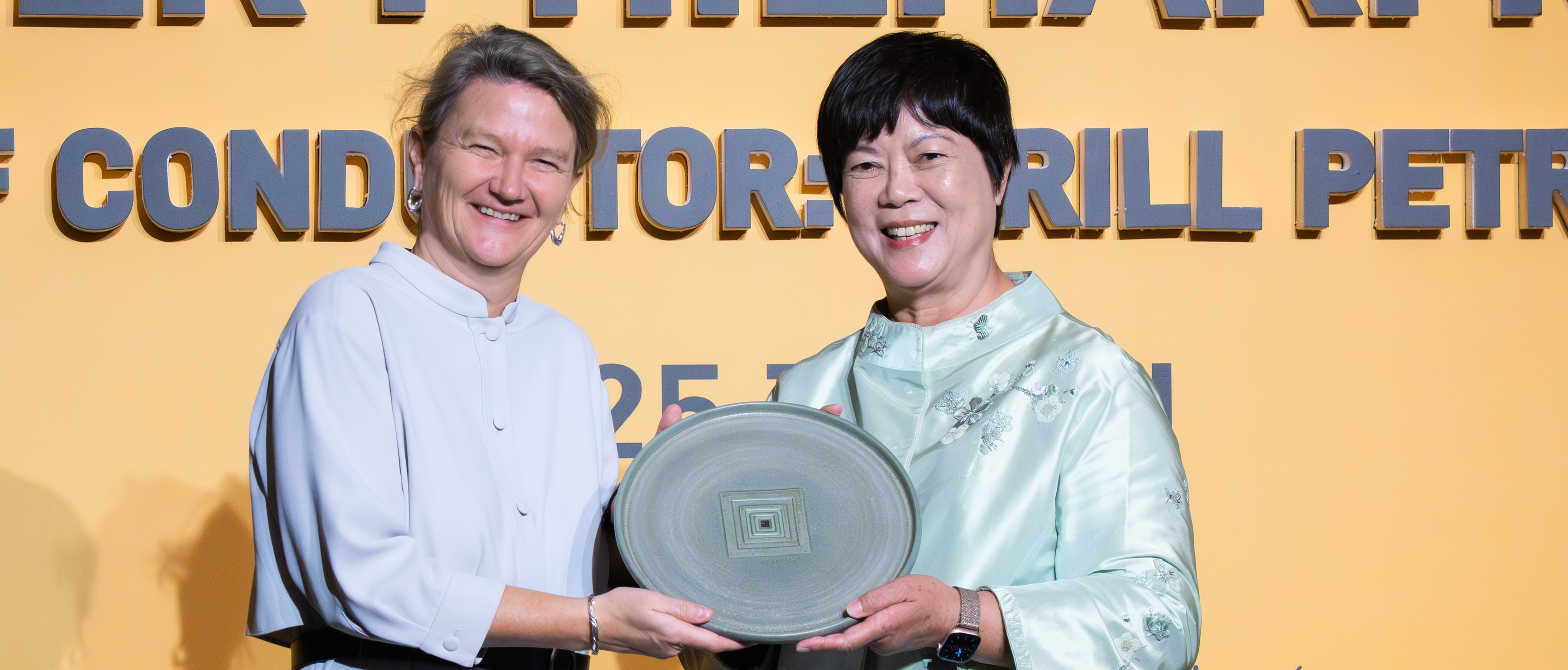 Zwei Frauen stehen lächelnd auf der Bühne vor einer gelben Kulisse. Die eine überreicht der anderen einen großen dekorativen Teller während einer offiziellen Veranstaltung oder Preisverleihung.