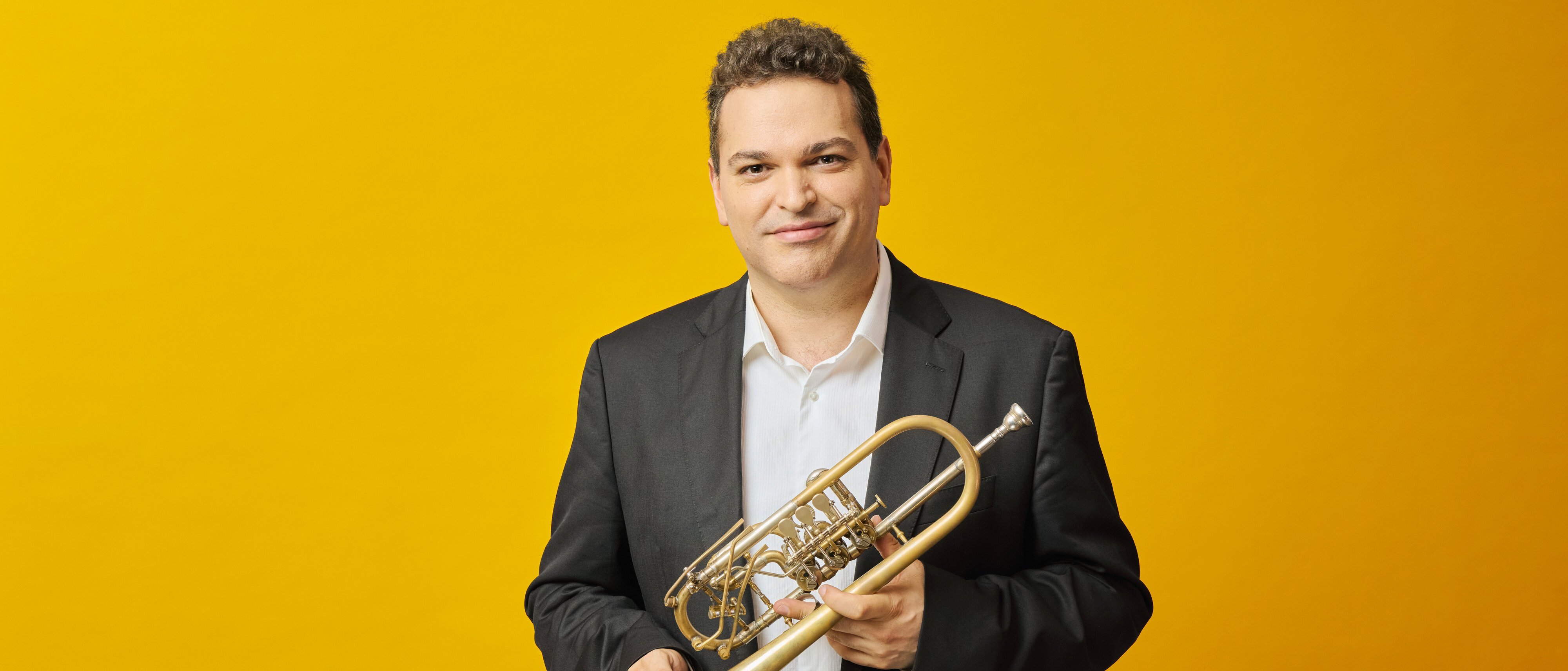 Ein Mann in schwarzem Anzug und weißem Hemd steht vor einem gelben Hintergrund, lächelt und hält eine Trompete.