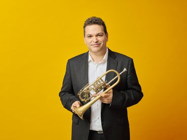 A man in a black suit and white shirt stands against a yellow background, holding a trumpet and smiling at the camera.