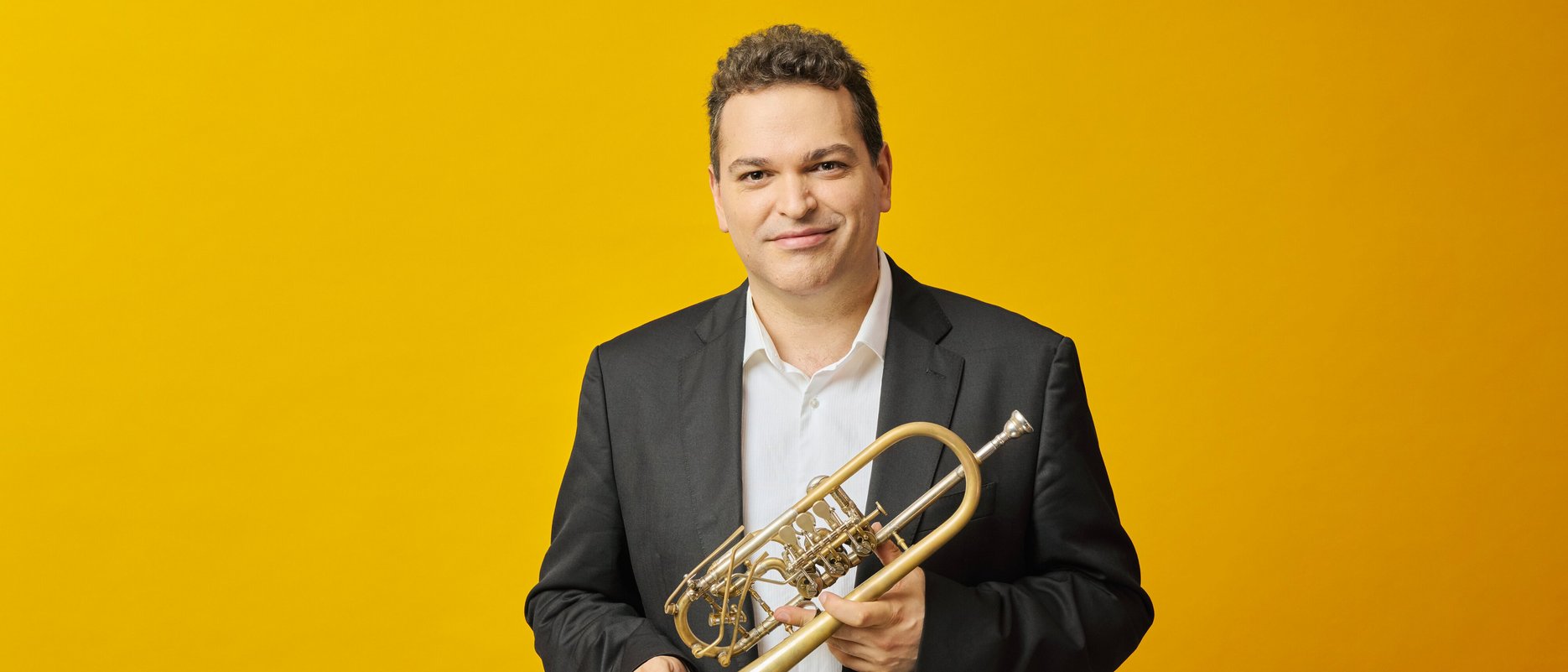 A man in a black suit and white shirt stands against a yellow background, holding a trumpet and smiling at the camera.