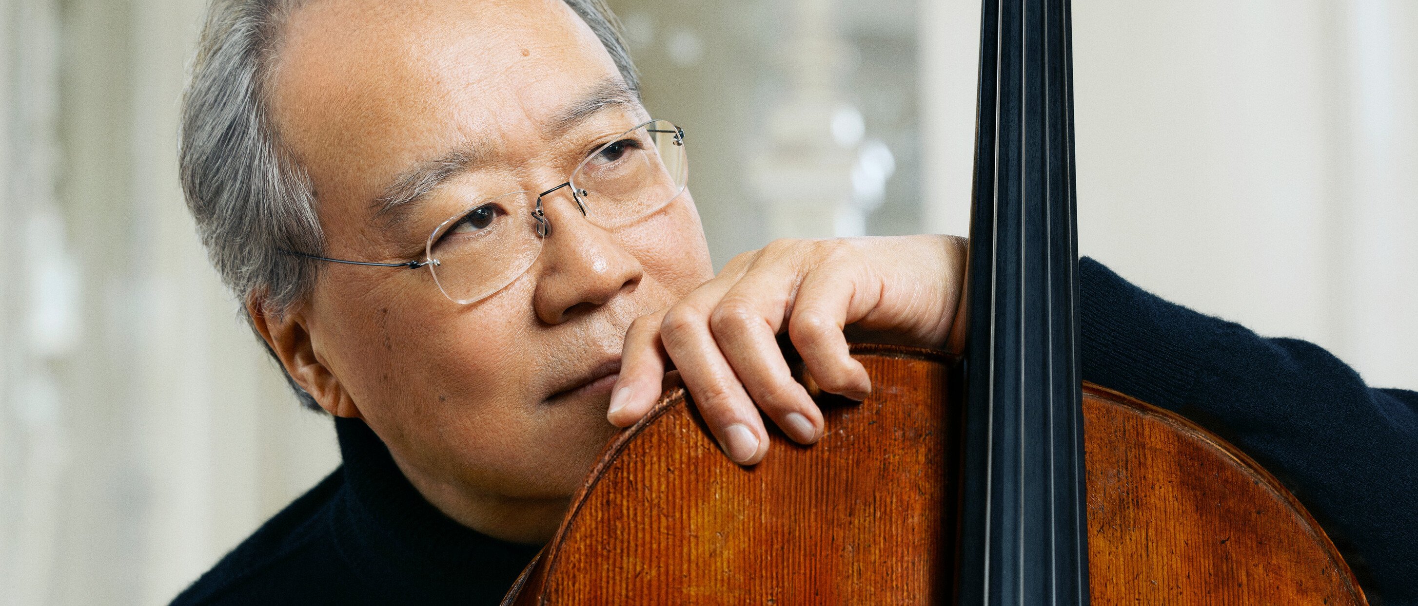 Yo-Yo Ma mit grauem Haar und Brille stützt sein Kinn und seine Hand auf ein Cello und schaut nachdenklich in die Ferne. Er trägt einen schwarzen Pullover und sitzt in einem sanft beleuchteten, eleganten Raum.