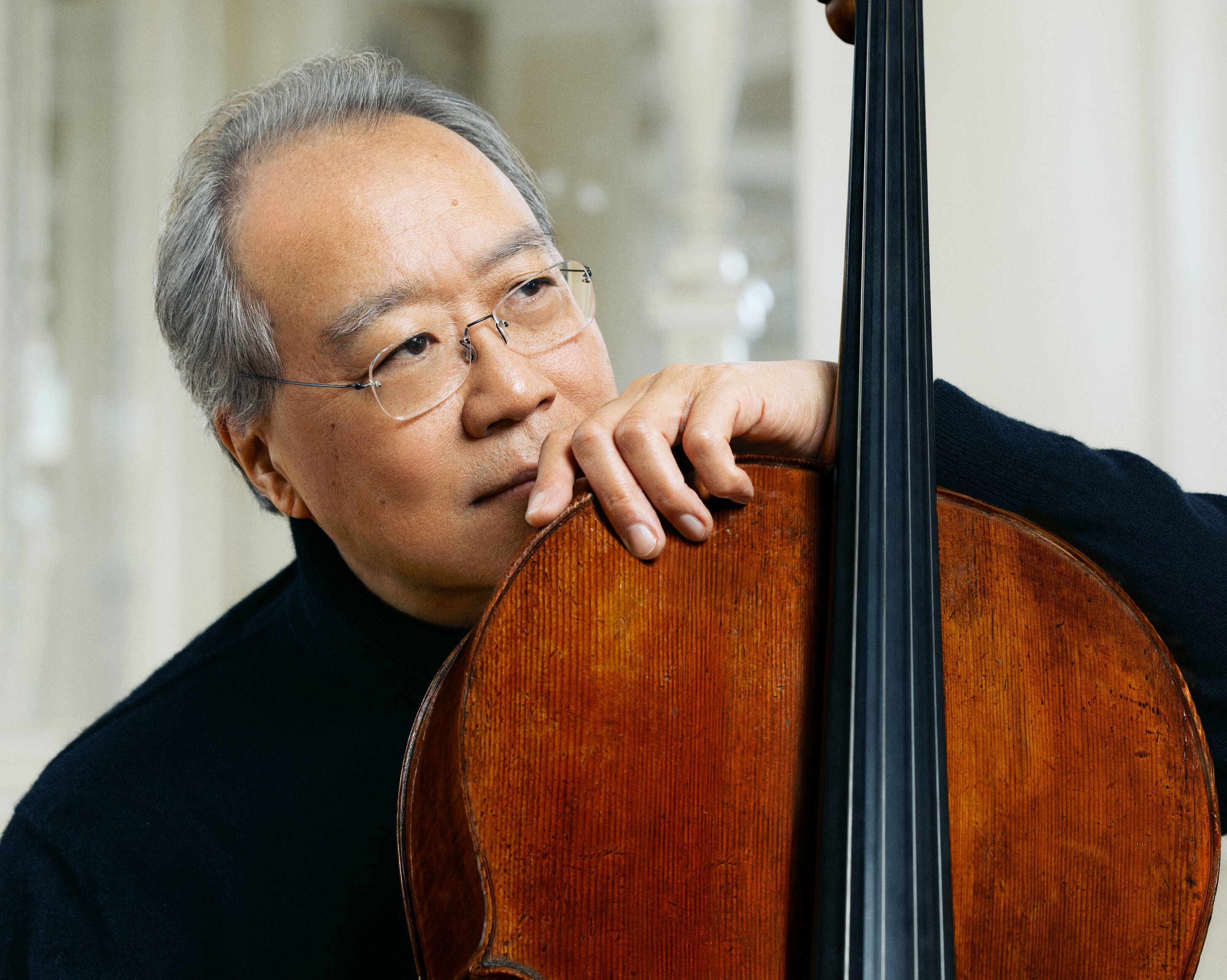 Yo-Yo Ma mit grauem Haar und Brille stützt sein Kinn und seine Hand auf ein Cello und schaut nachdenklich in die Ferne. Er trägt einen schwarzen Pullover und sitzt in einem sanft beleuchteten, eleganten Raum.