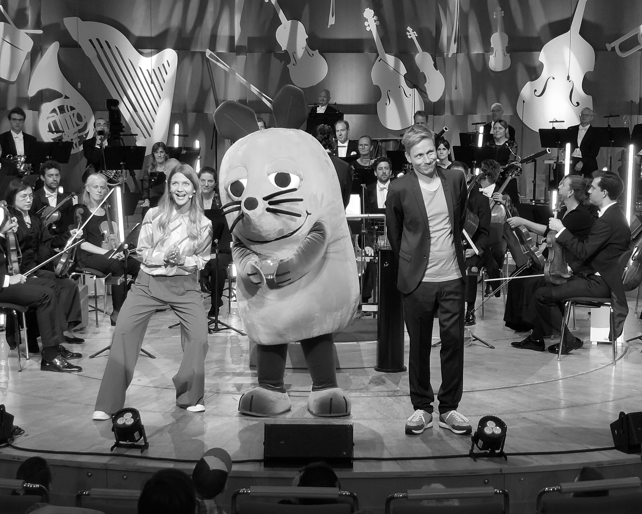 Ein großes Orchester spielt auf der Bühne, auf der zwei lächelnde Moderatoren und eine Person in einem großen Mauskostüm stehen, während das Publikum zuschaut.