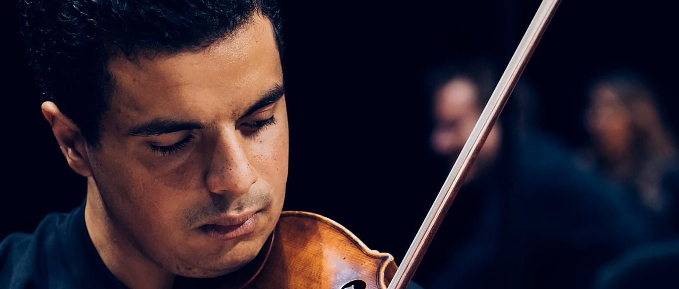 Ein Mann mit kurzen, dunklen Haaren spielt konzentriert Geige, wobei er das Instrument an der Schulter hält und den Bogen führt; im Hintergrund ist ein dunkles, verschwommenes Bild zu sehen.