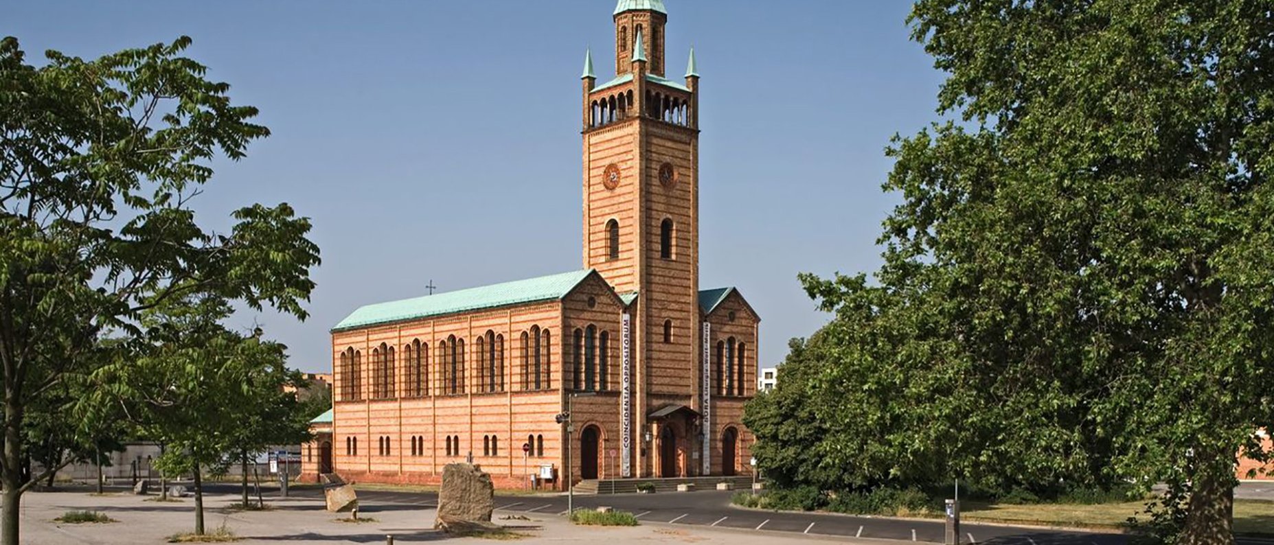 A brick church with a tall green spire and arched windows stands in an open area, surrounded by trees and an empty parking lot under a clear blue sky.