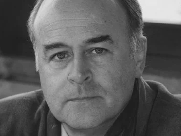 Ein Mann mittleren Alters mit Glatze und ernster Miene blickt in diesem Schwarz-Weiß-Porträt in Großaufnahme direkt in die Kamera. Er trägt eine Jacke und sitzt in einem Innenraum in der Nähe eines Fensters.