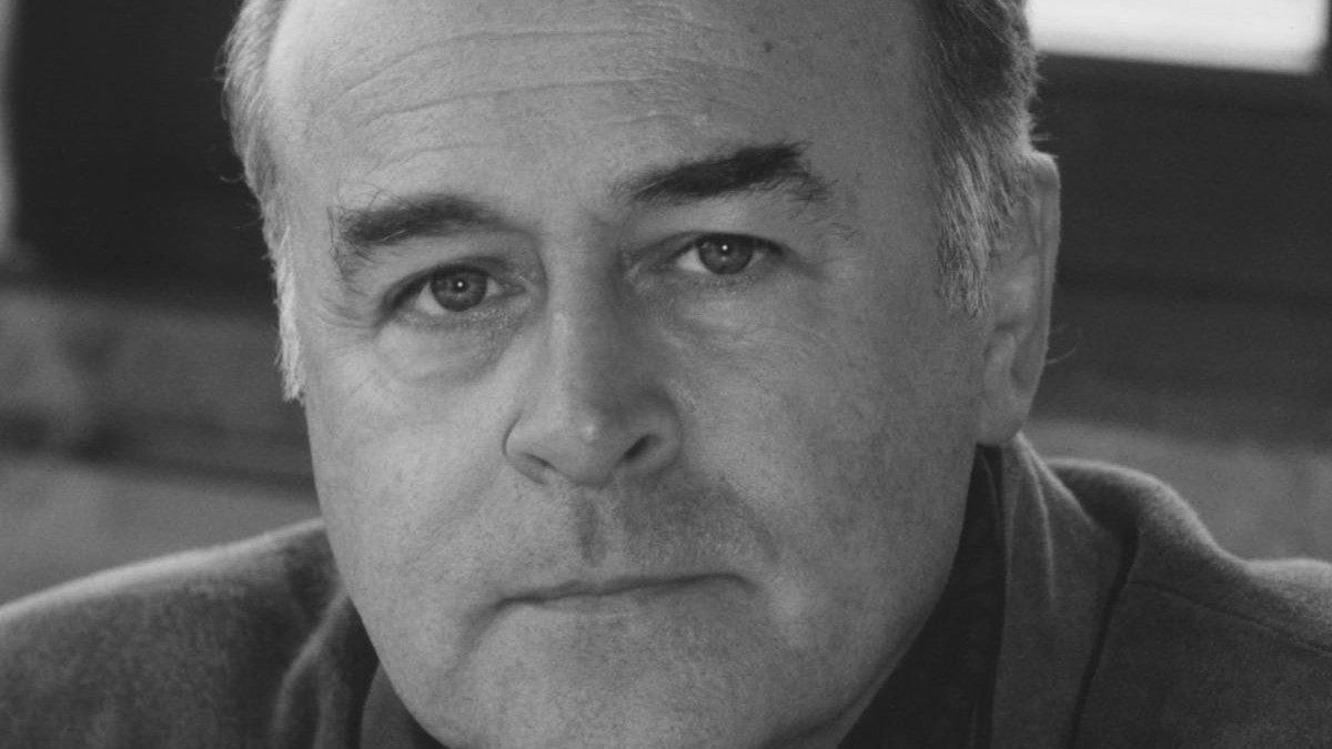 Ein Mann mittleren Alters mit Glatze und ernster Miene blickt in diesem Schwarz-Weiß-Porträt in Großaufnahme direkt in die Kamera. Er trägt eine Jacke und sitzt in einem Innenraum in der Nähe eines Fensters.