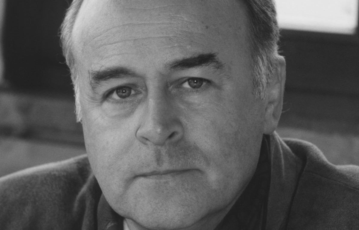 A black and white close-up photo of an older man with a receding hairline, looking directly at the camera with a neutral expression. He is indoors, wearing a collared shirt and jacket.