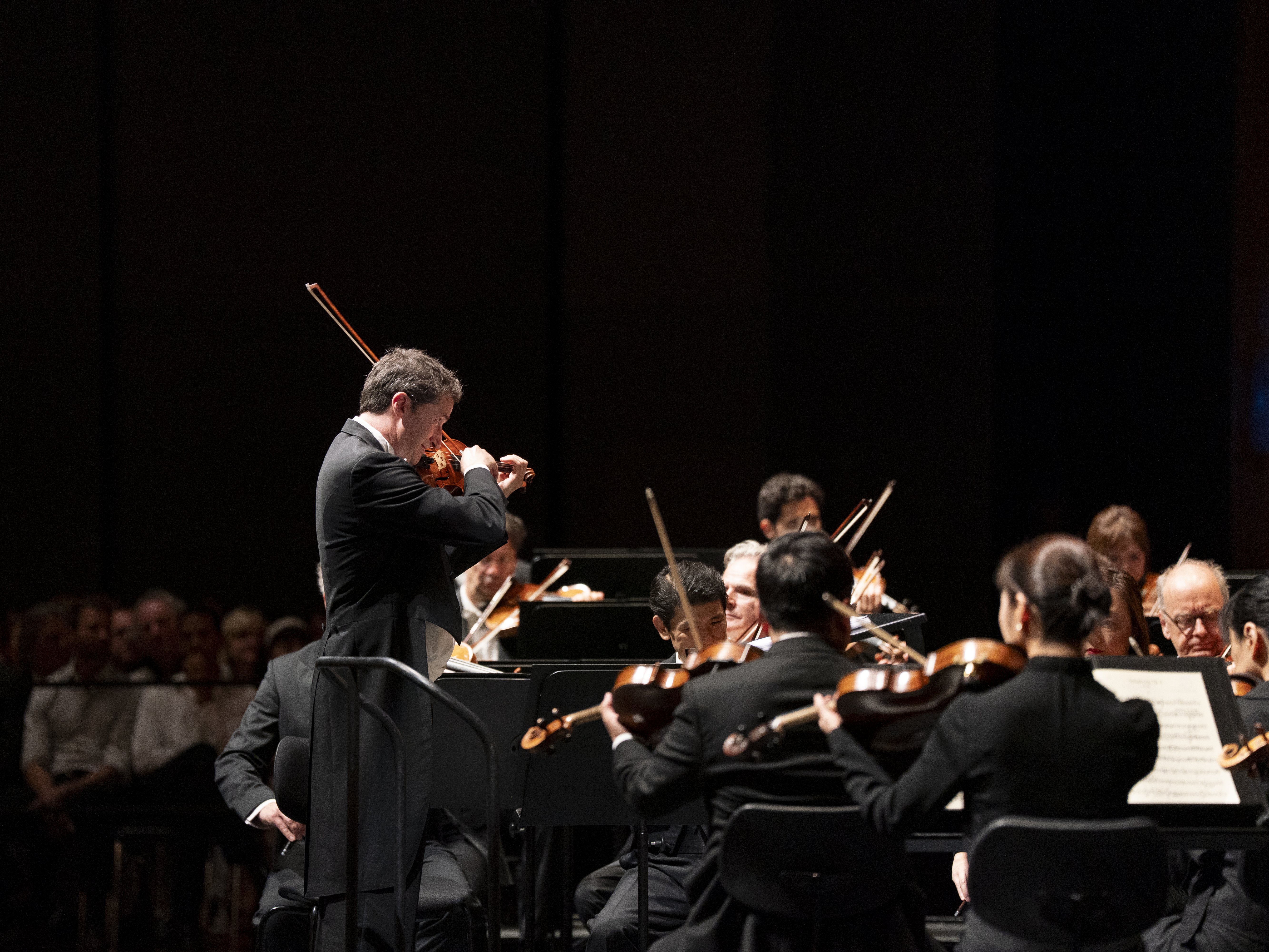 A violinist stands and performs with an orchestra on stage in formal attire, while an audience watches attentively in the background.