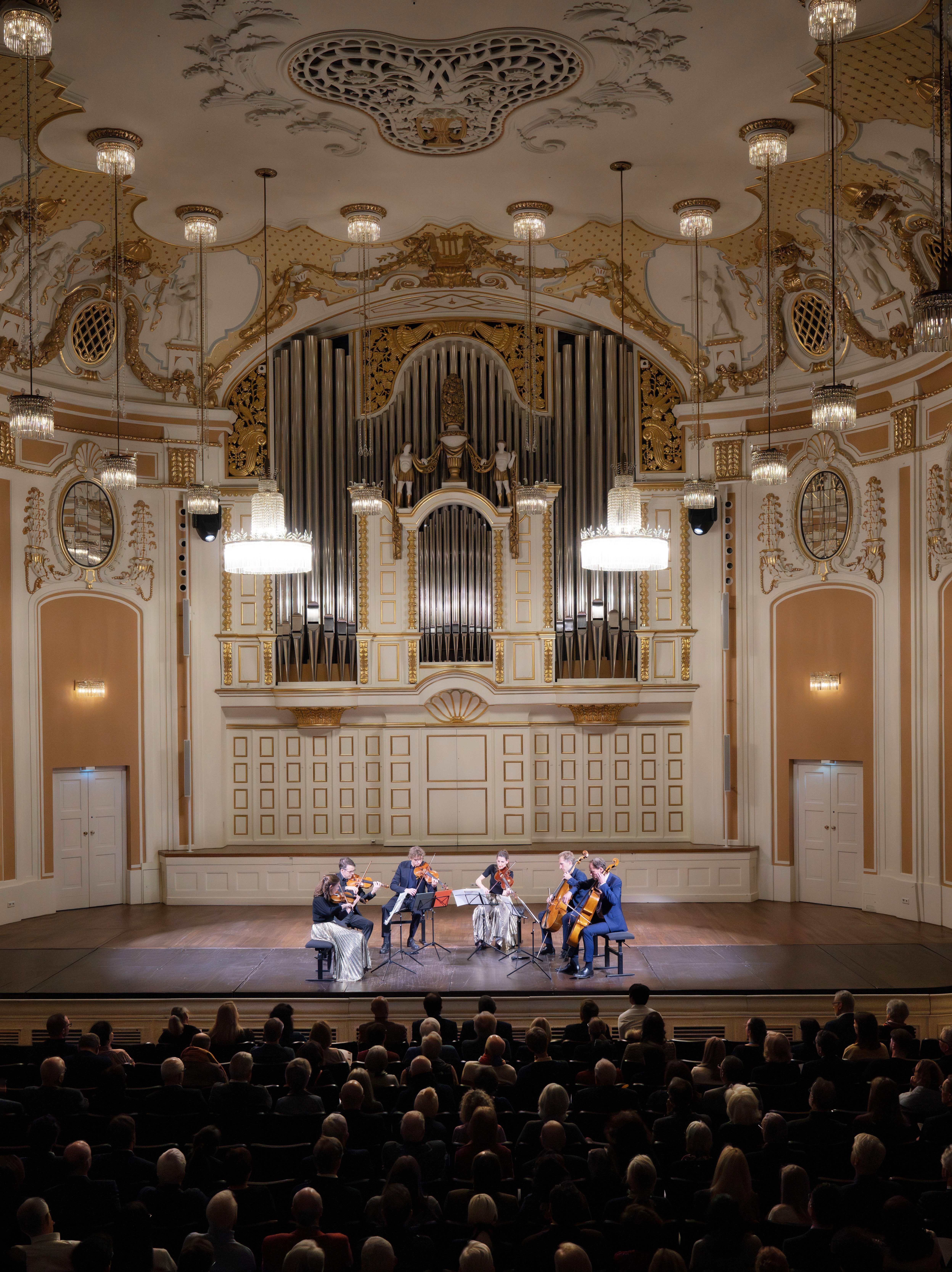 Ein Streichquintett spielt auf der Bühne eines prunkvollen Konzertsaals mit einer großen Pfeifenorgel, Kronleuchtern und einem Publikum, das vor den Musikern sitzt.