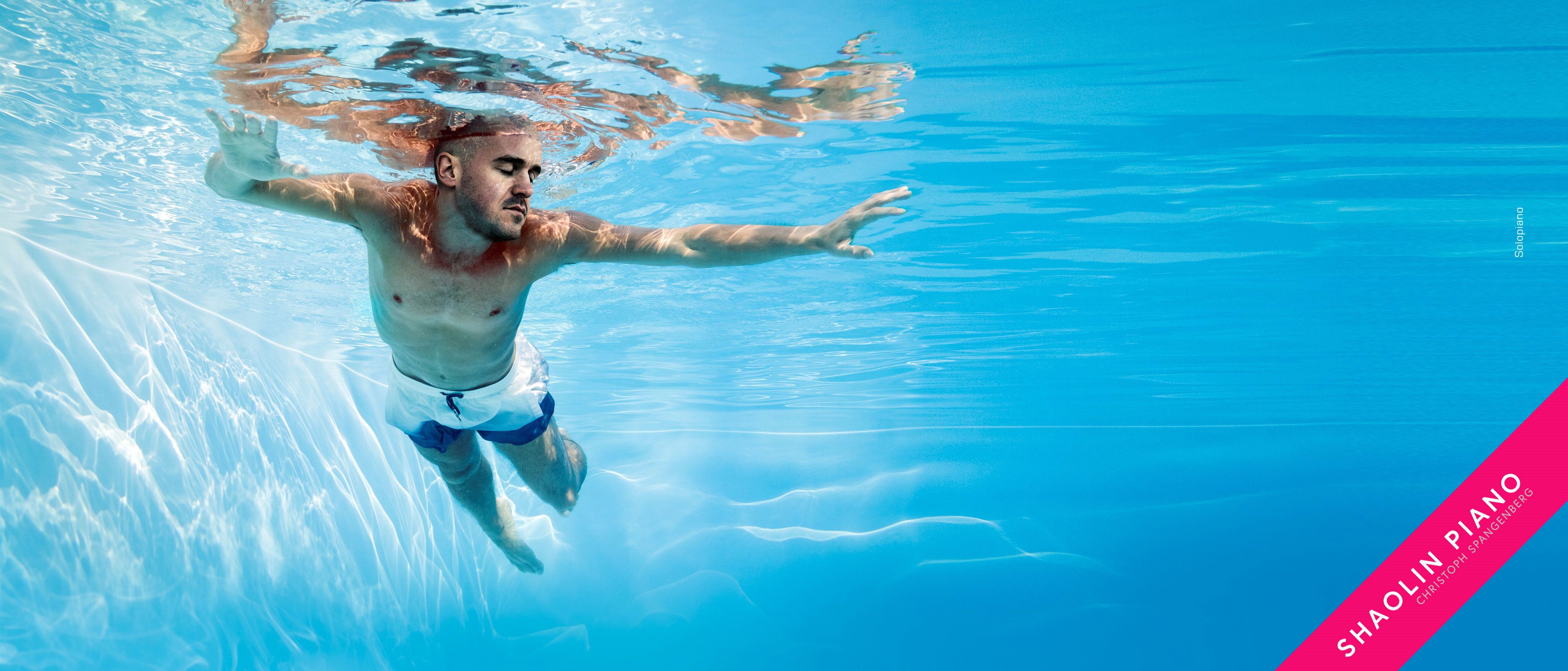 A man swims underwater in a pool, wearing white swim trunks. Sunlight creates patterns on the pool floor. The words Shaolin Piano appear in white text on a pink diagonal stripe in the corner.