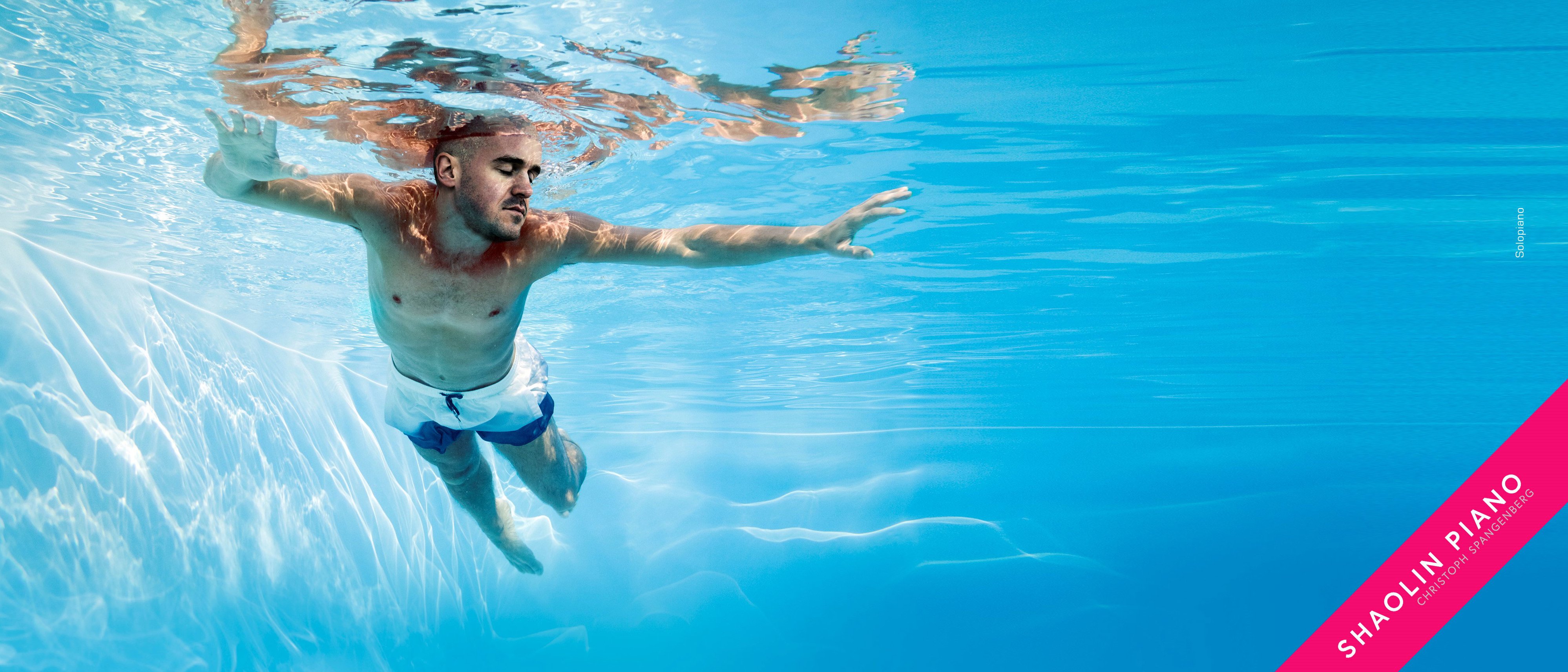 Ein Mann in Badehose treibt unter Wasser in einem blauen Schwimmbecken und streckt einen Arm nach vorne aus. Sonnenlicht fällt durch das Wasser. Auf einem rosa diagonalen Streifen ist der Schriftzug „Shaolin Piano» zu sehen.