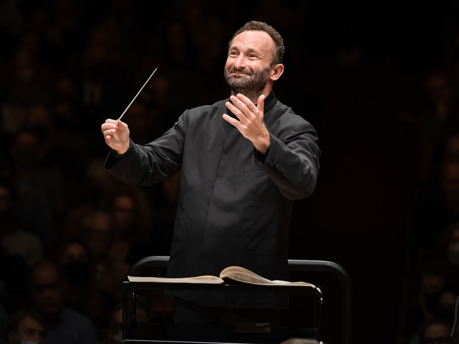 Kirill Petrenko conducts the orchestra with a baton. He stands on a podium, with sheet music in front of him. Wearing a black suit, he has a focused expression, and in the background, there is a dimly lit audience.