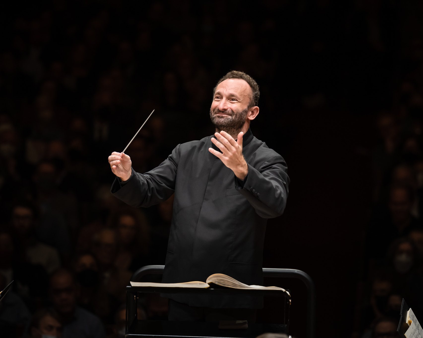 Kirill Petrenko mit einem Taktstock leitet das Orchester. Er steht auf einem Podium, vor ihm liegen Notenblätter. Er trägt einen schwarzen Anzug, hat einen konzentrierten Gesichtsausdruck und im Hintergrund ein schwach beleuchtetes Publikum.
