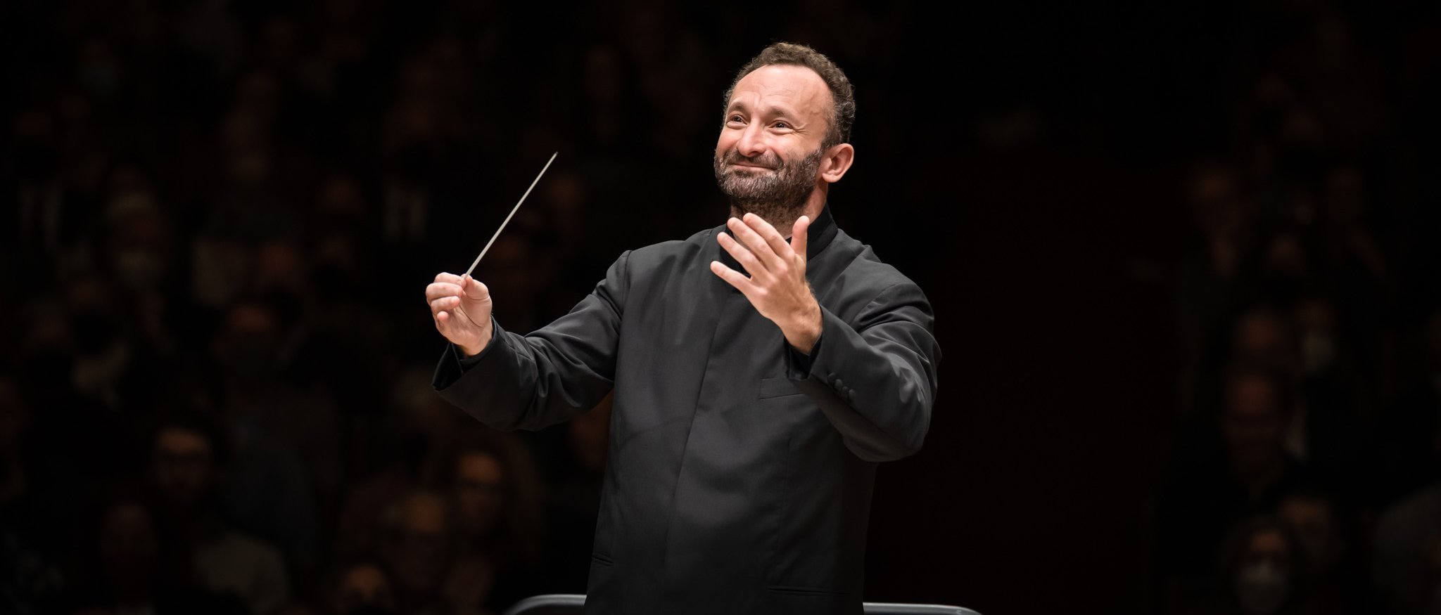 Kirill Petrenko mit einem Taktstock leitet das Orchester. Er steht auf einem Podium, vor ihm liegen Notenblätter. Er trägt einen schwarzen Anzug, hat einen konzentrierten Gesichtsausdruck und im Hintergrund ein schwach beleuchtetes Publikum.