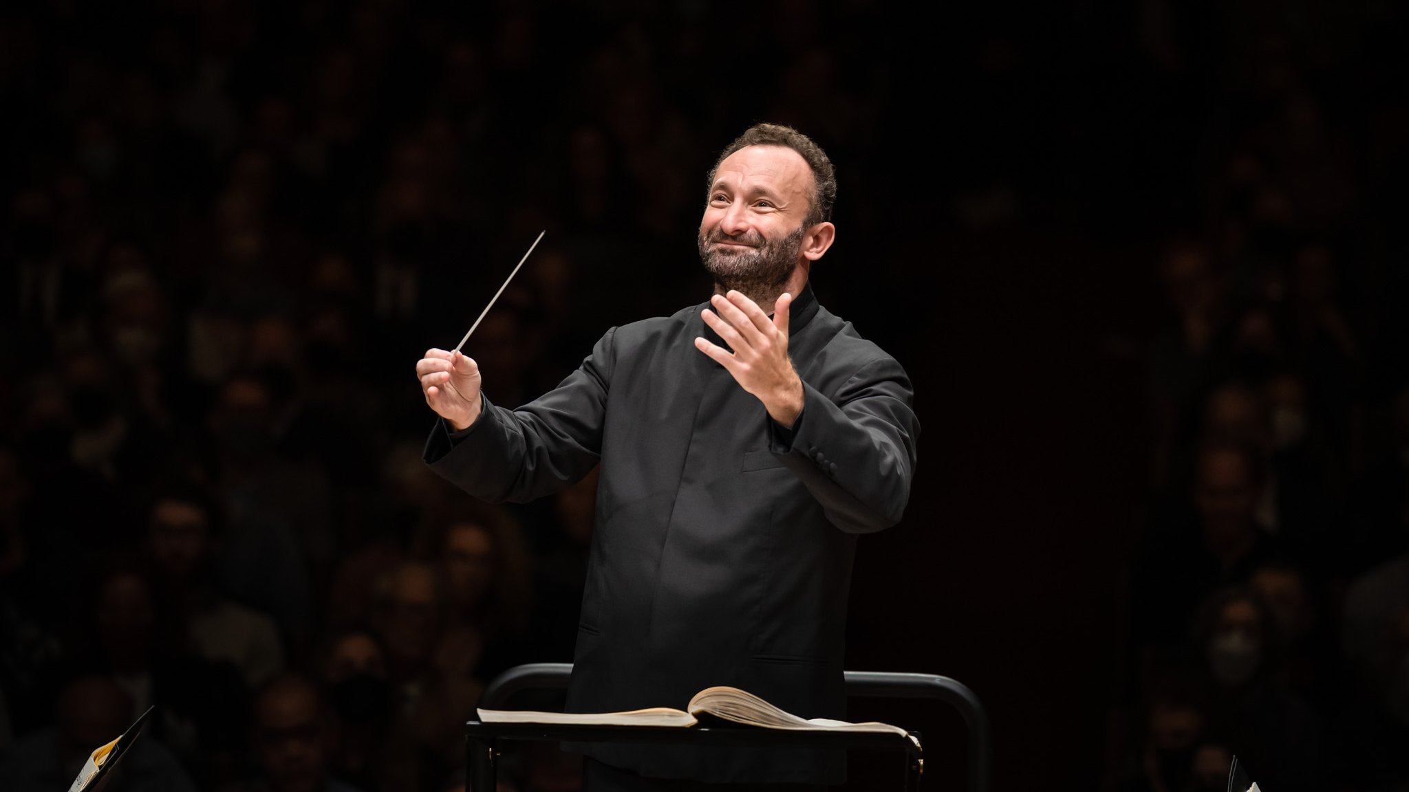 Kirill Petrenko mit einem Taktstock leitet das Orchester. Er steht auf einem Podium, vor ihm liegen Notenblätter. Er trägt einen schwarzen Anzug, hat einen konzentrierten Gesichtsausdruck und im Hintergrund ein schwach beleuchtetes Publikum.