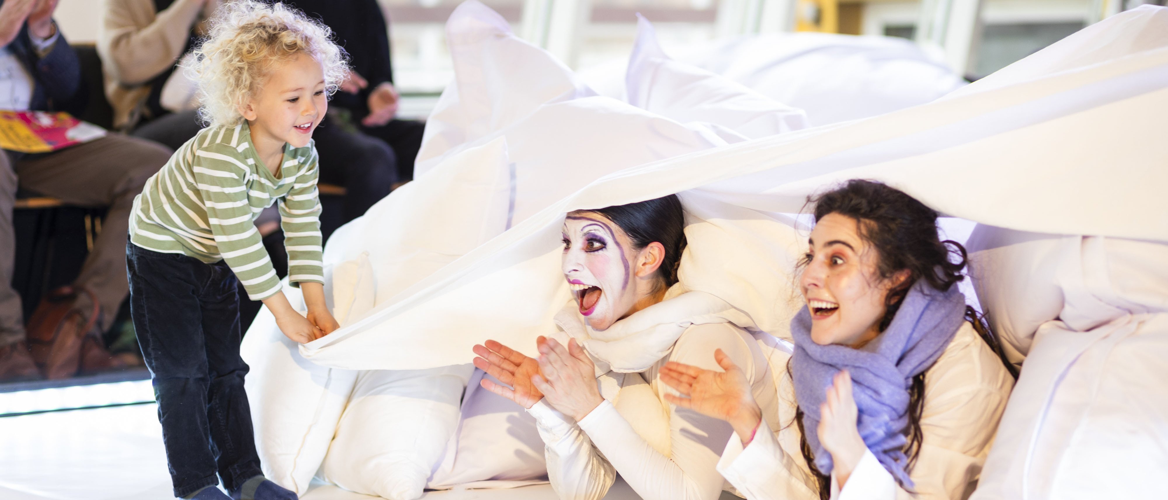 Child lifts up a blanket, two women with colourful make-up lie underneath and look surprised