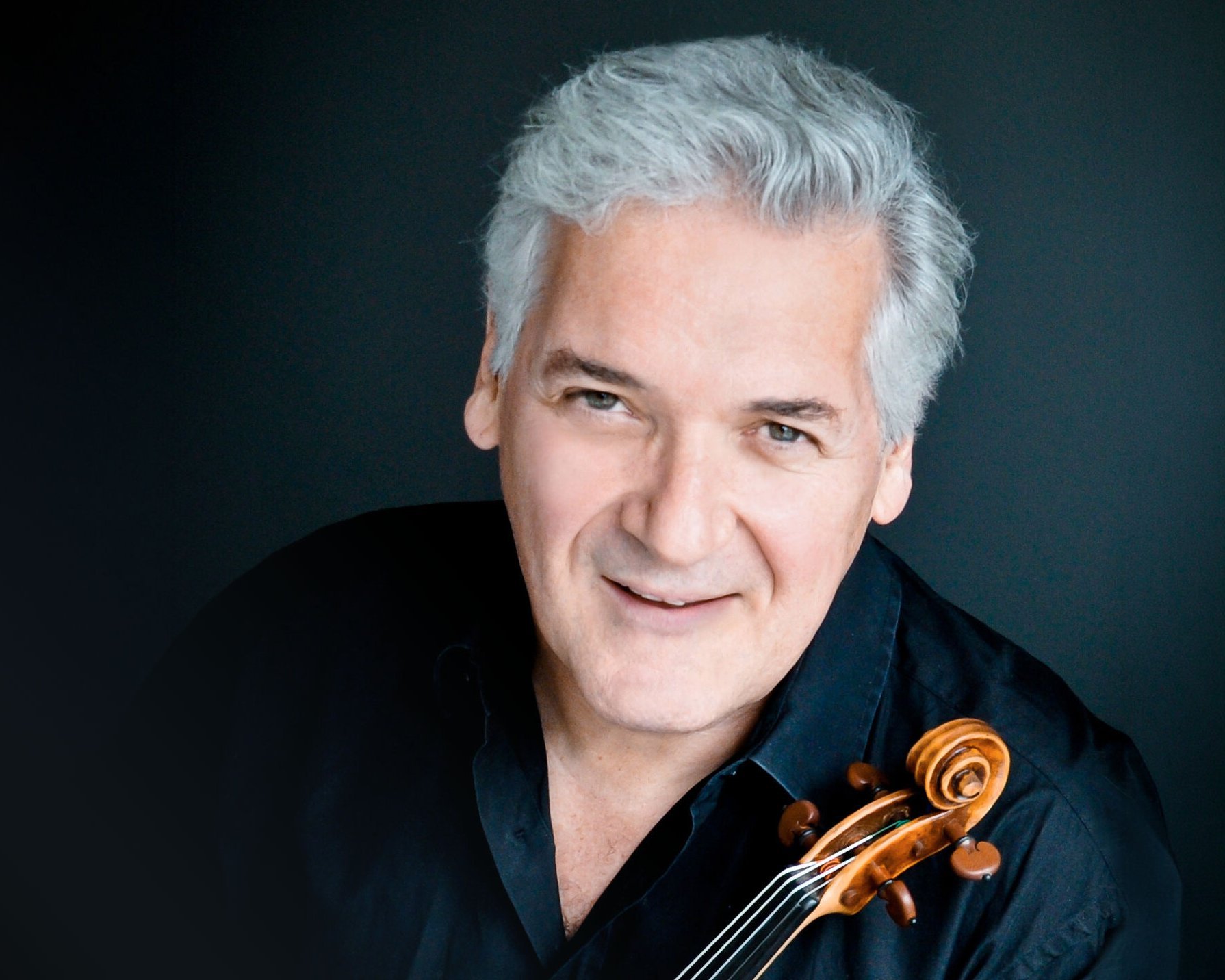 Ein lächelnder Pinchas Zukerman mit grauem Haar und schwarzem Hemd posiert mit einer Geige vor einem dunklen Hintergrund.