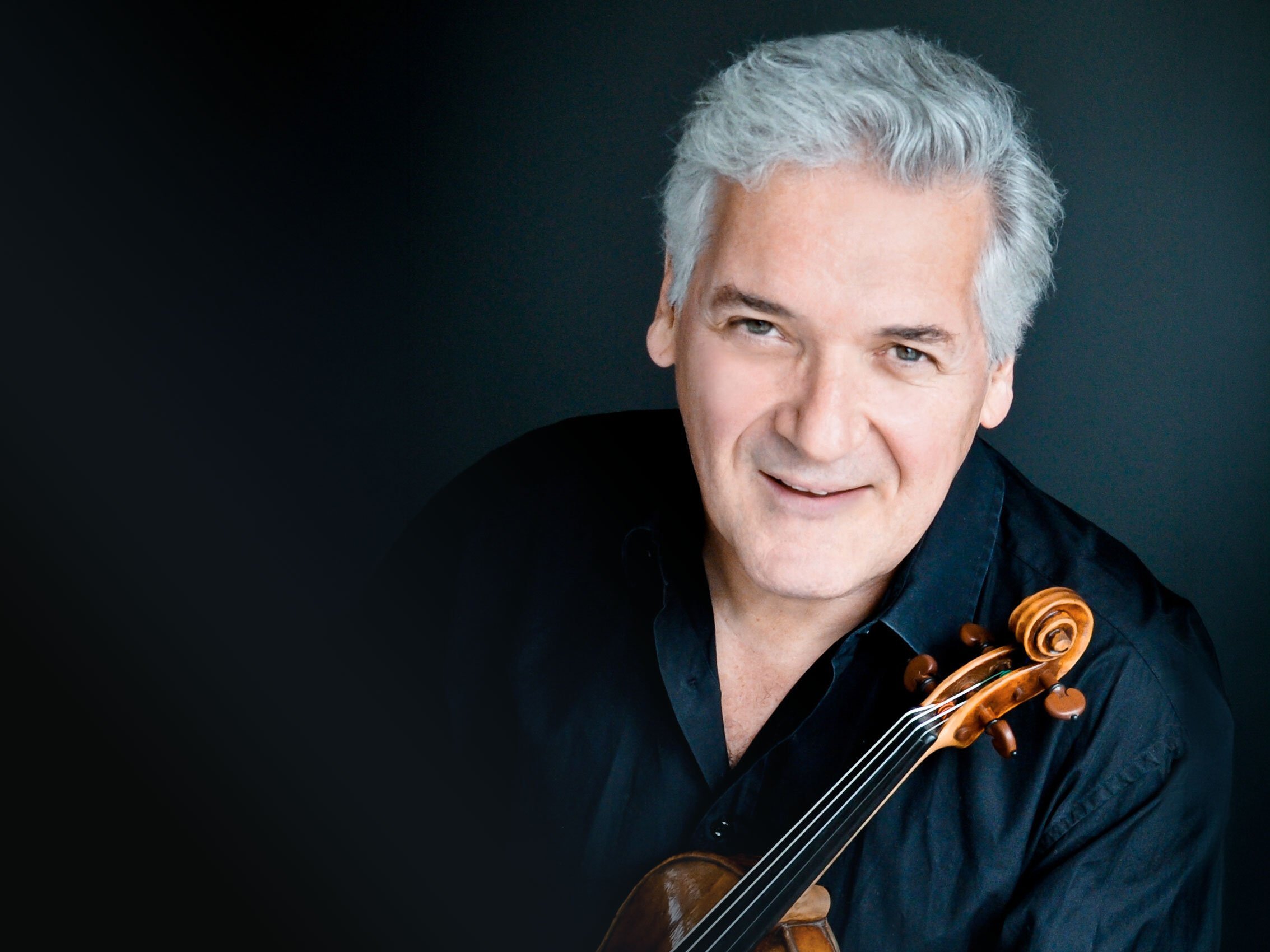 Ein lächelnder Pinchas Zukerman mit grauem Haar und schwarzem Hemd posiert mit einer Geige vor einem dunklen Hintergrund.