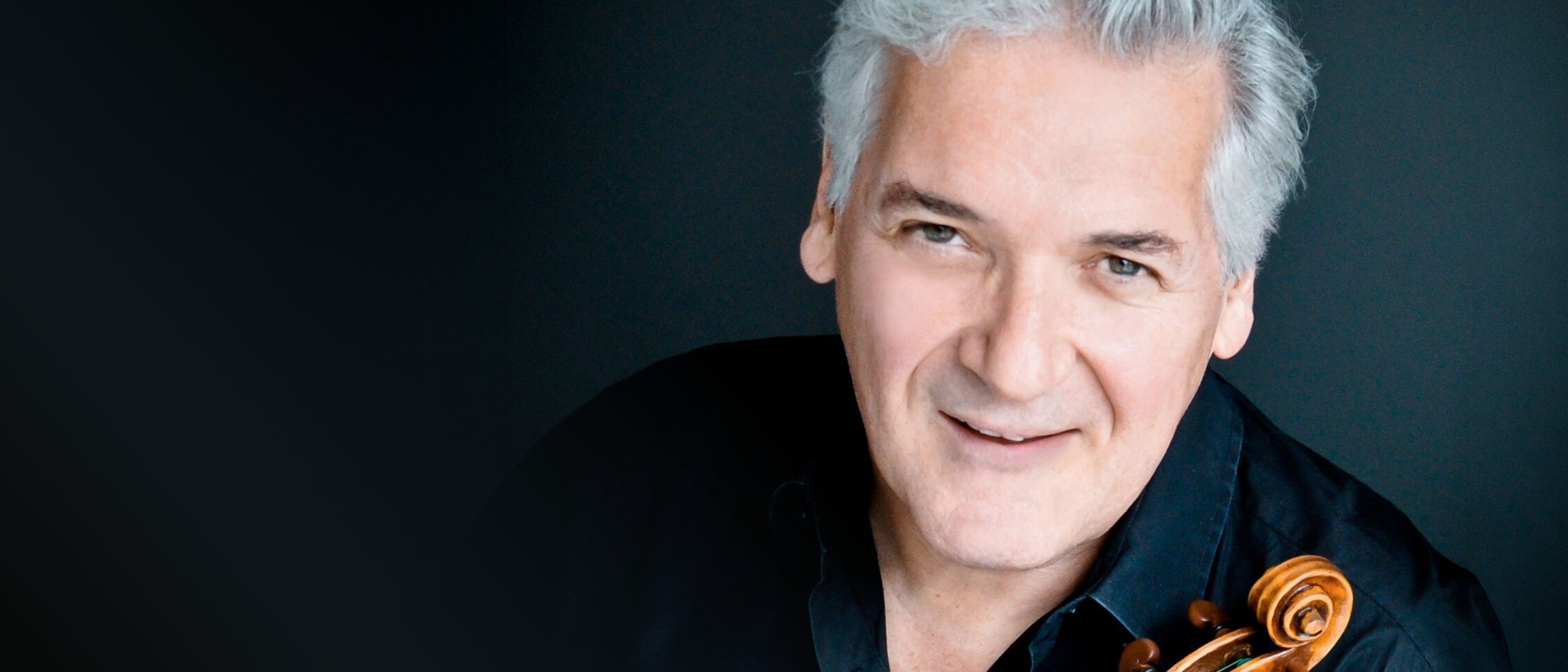 Ein lächelnder Pinchas Zukerman mit grauem Haar und schwarzem Hemd posiert mit einer Geige vor einem dunklen Hintergrund.