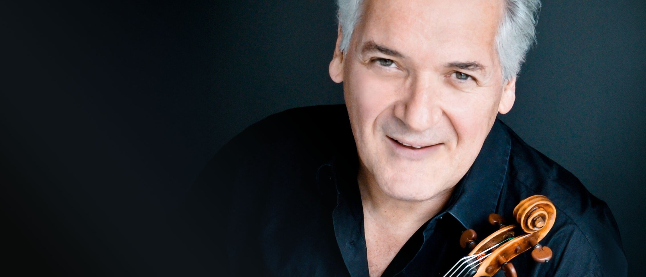 Ein lächelnder Pinchas Zukerman mit grauem Haar und schwarzem Hemd posiert mit einer Geige vor einem dunklen Hintergrund.