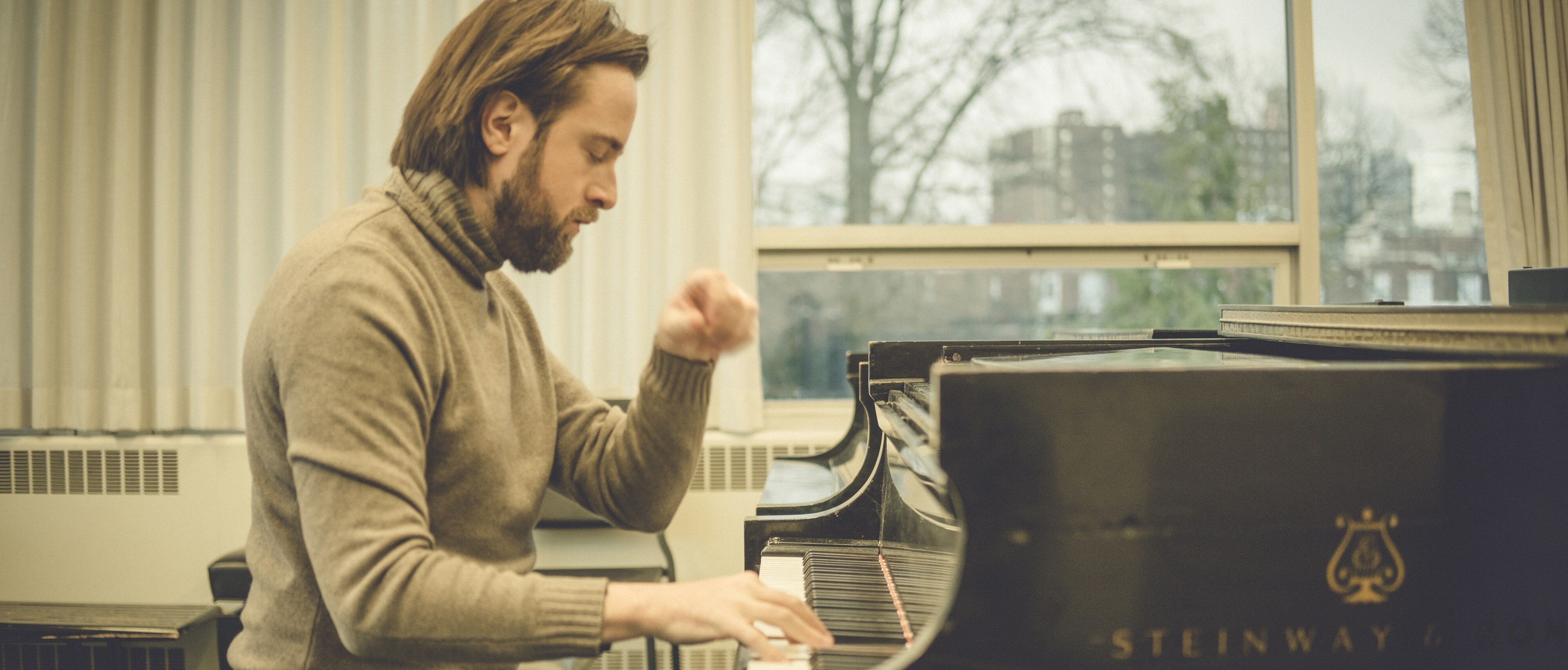 Daniil Trifonov mit schulterlangem Haar und Bart spielt in einem hellen Raum mit großen Fenstern auf einem Steinway & Sons-Flügel, er trägt einen grauen Rollkragenpullover und Jeans.