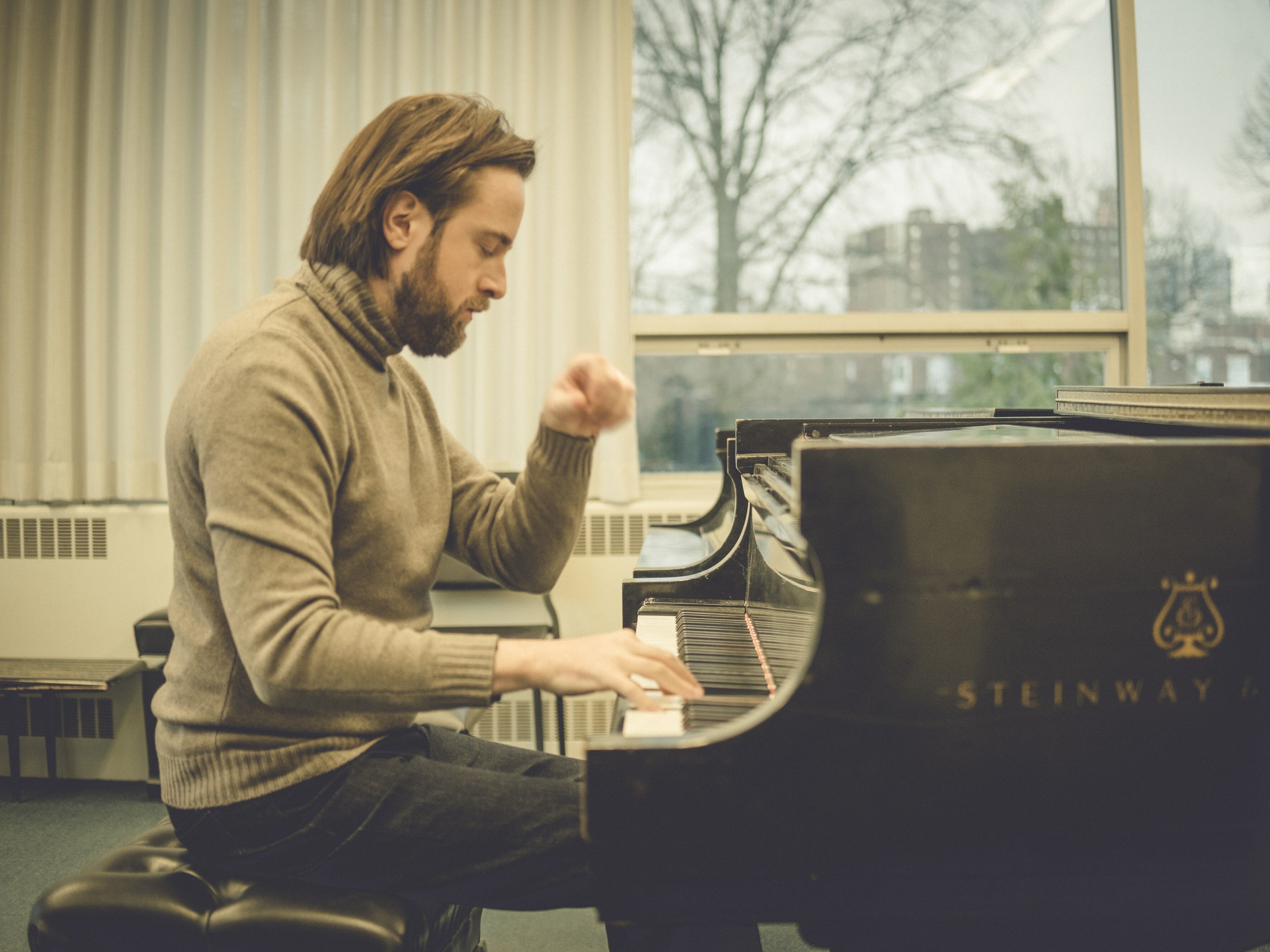Daniil Trifonov with shoulder-length hair and a beard plays a grand piano in a well-lit room with large windows, wearing a gray turtleneck sweater and jeans. The piano is a Steinway & Sons.