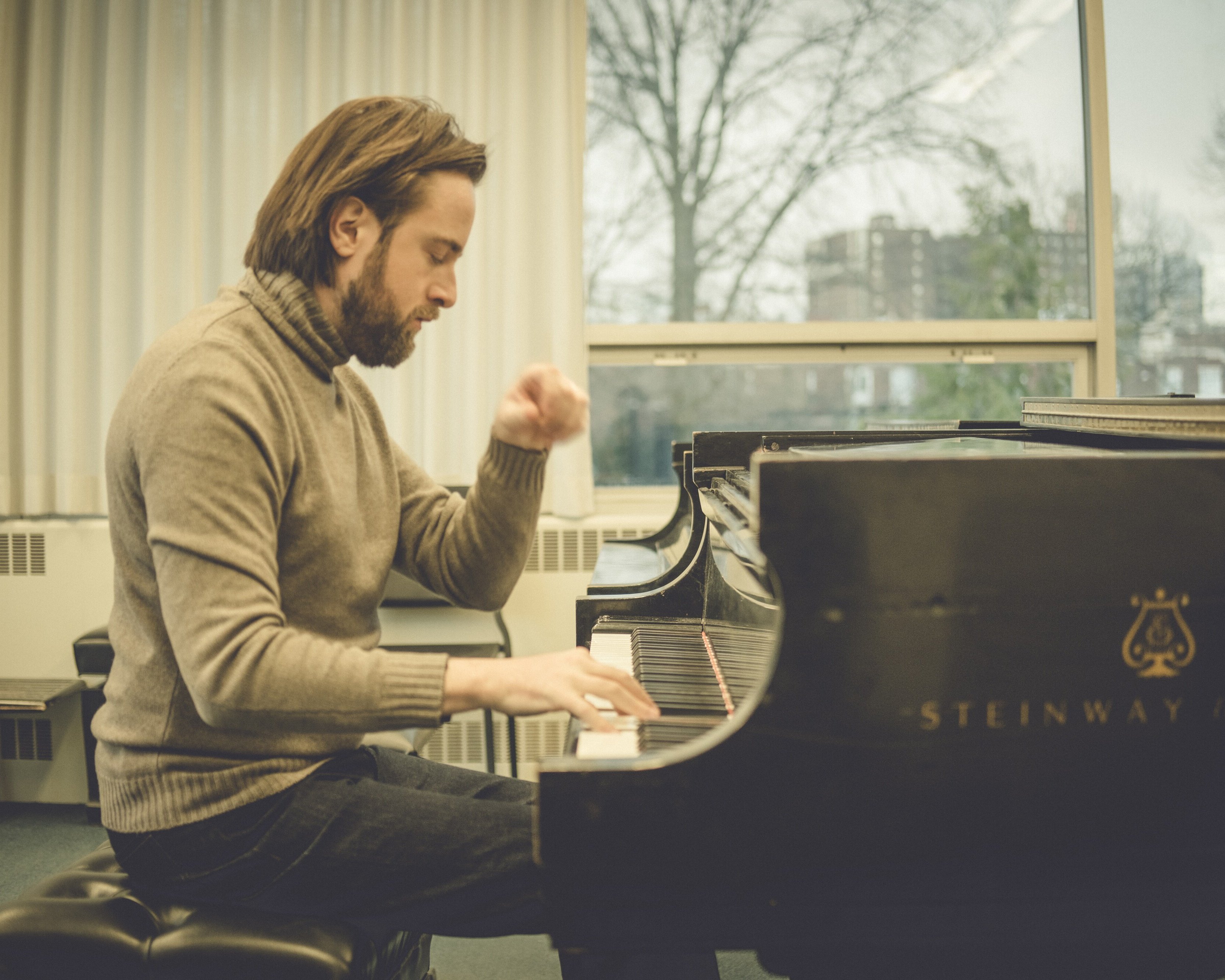 Daniil Trifonov mit schulterlangem Haar und Bart spielt in einem hellen Raum mit großen Fenstern auf einem Steinway & Sons-Flügel, er trägt einen grauen Rollkragenpullover und Jeans.