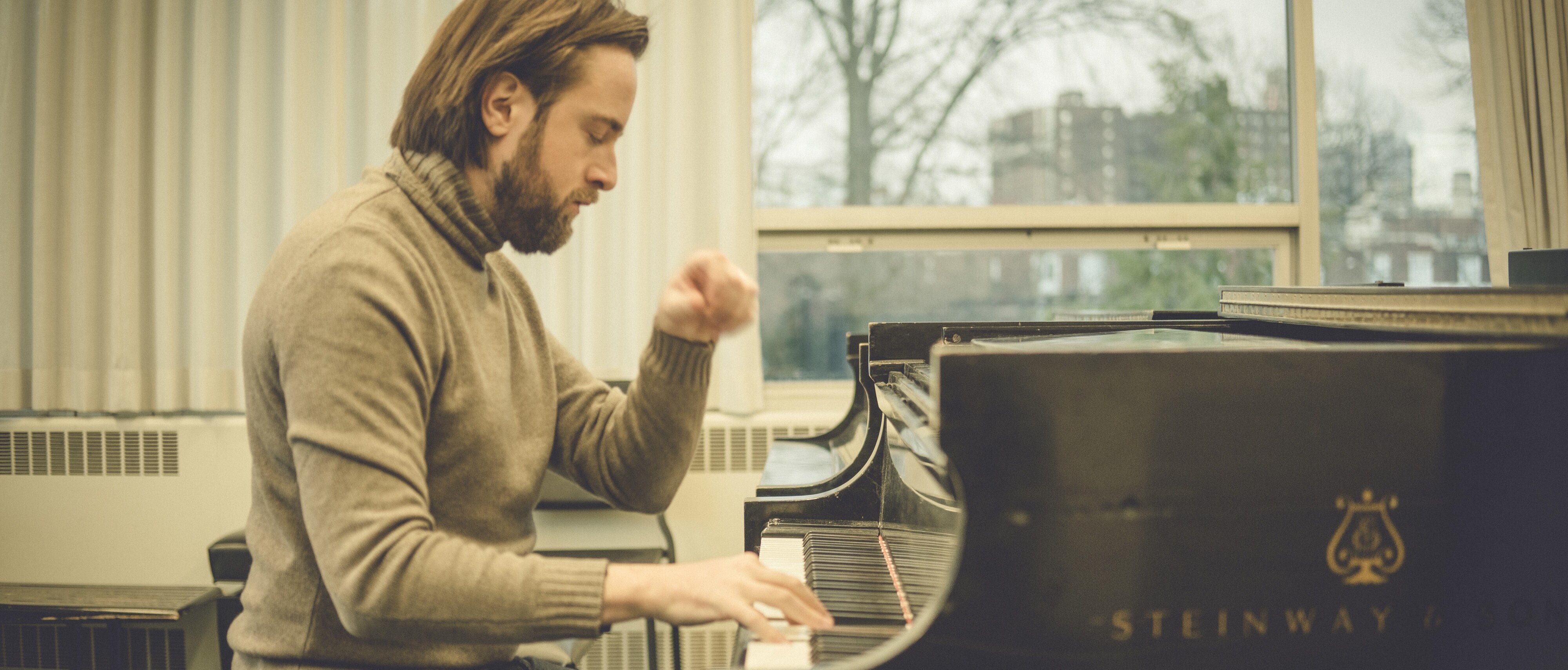 Daniil Trifonov mit schulterlangem Haar und Bart spielt in einem hellen Raum mit großen Fenstern auf einem Steinway & Sons-Flügel, er trägt einen grauen Rollkragenpullover und Jeans.