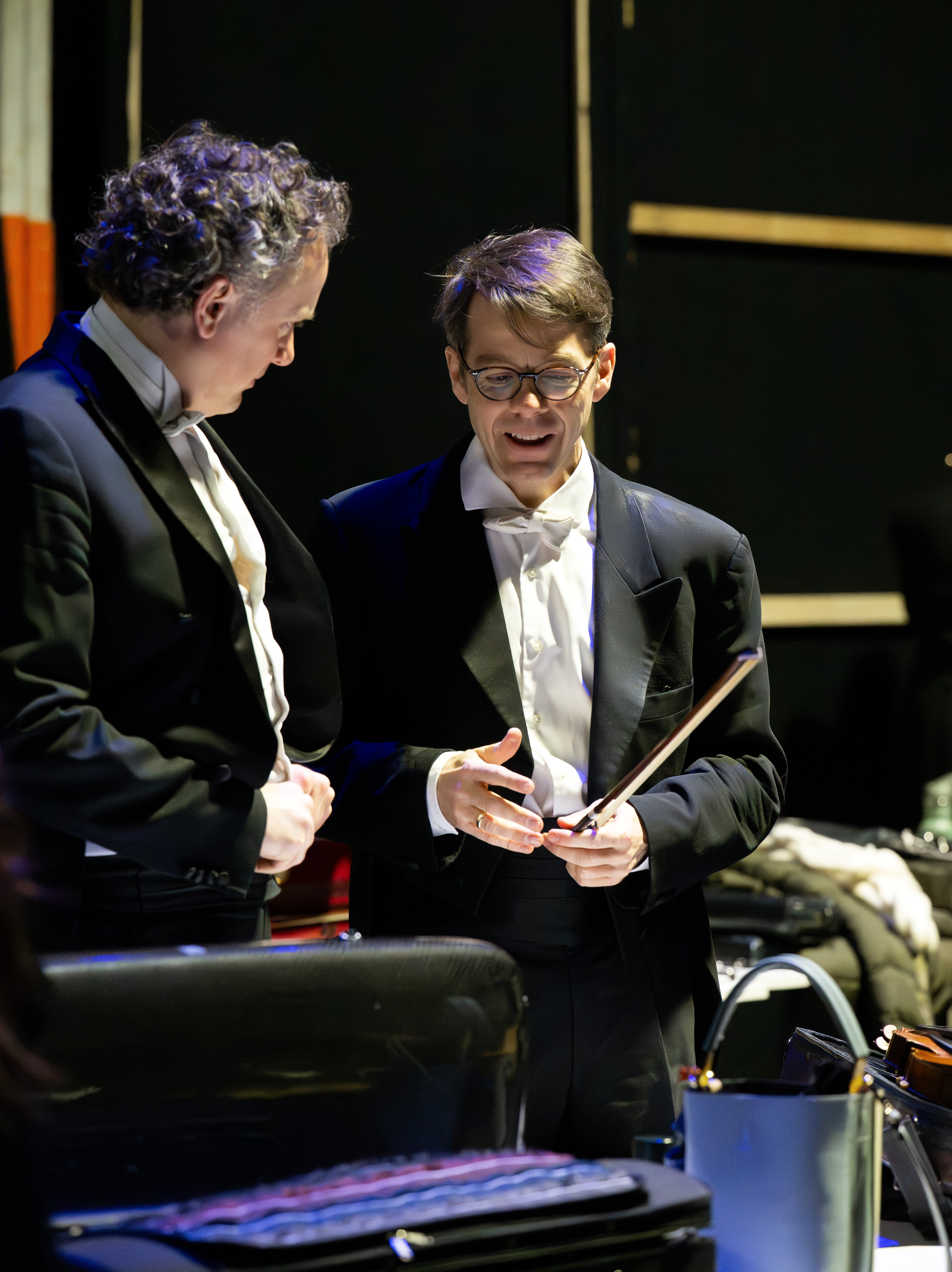 Zwei Männer in formellen Smokings stehen hinter der Bühne und führen ein Gespräch. Einer hält einen Dirigentenstab, und beide wirken konzentriert. Im Vordergrund sind Musikinstrumente und Koffer zu sehen.