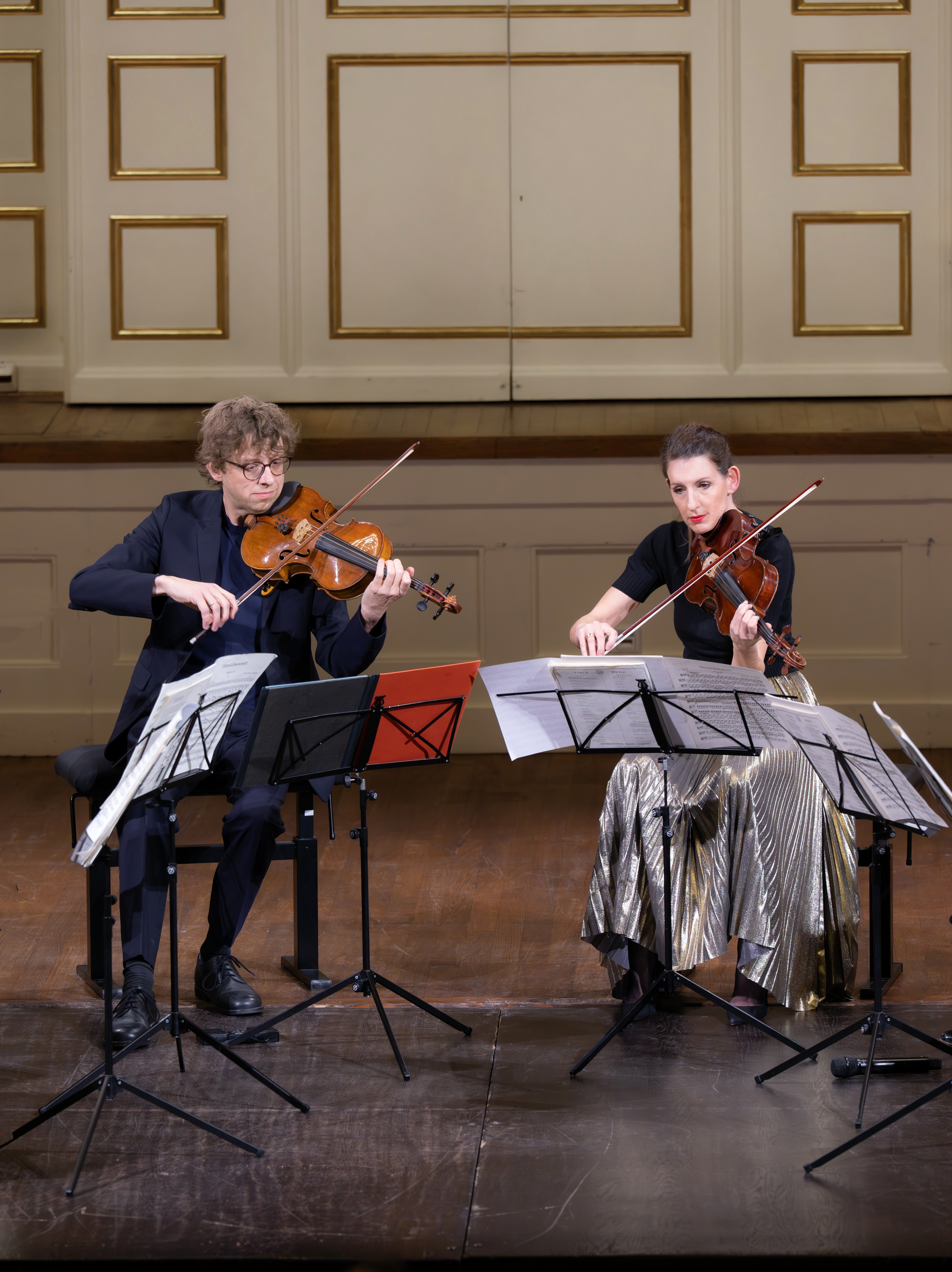 Zwei Musiker, ein Mann und eine Frau, sitzen auf der Bühne und spielen Geige. Sie lesen die Noten von Ständern ab und tragen formelle schwarze Kleidung; die Frau trägt einen silberglänzenden Rock. Der Hintergrund ist eine verzierte Wand.