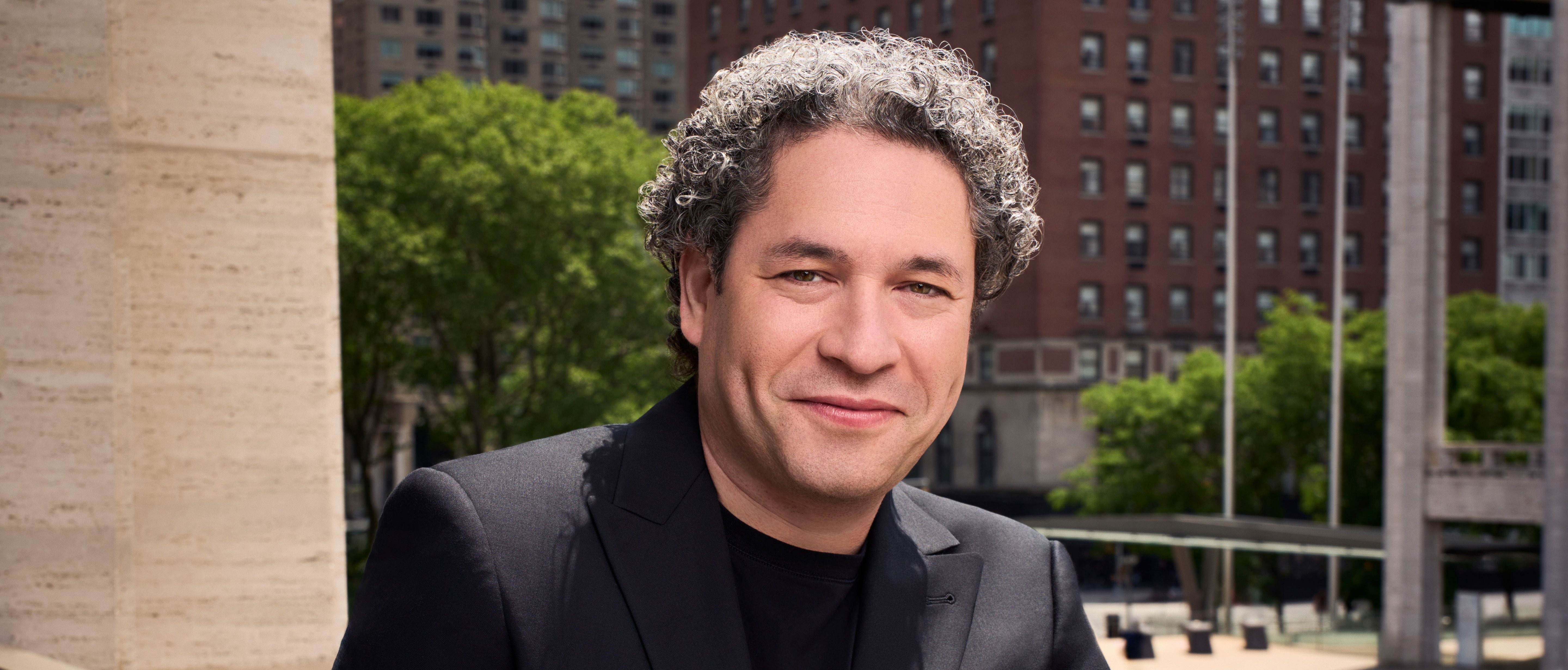 A man with curly gray hair, wearing a black blazer and shirt, smiles while standing outside in front of tall buildings and green trees on a sunny day.