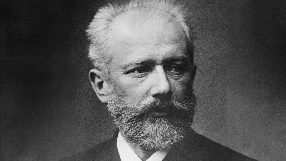 Black and white portrait of Pyotr Tchaikovsky with a full beard and mustache, short light hair, wearing a suit and tie, gazing slightly to his left with a serious expression.