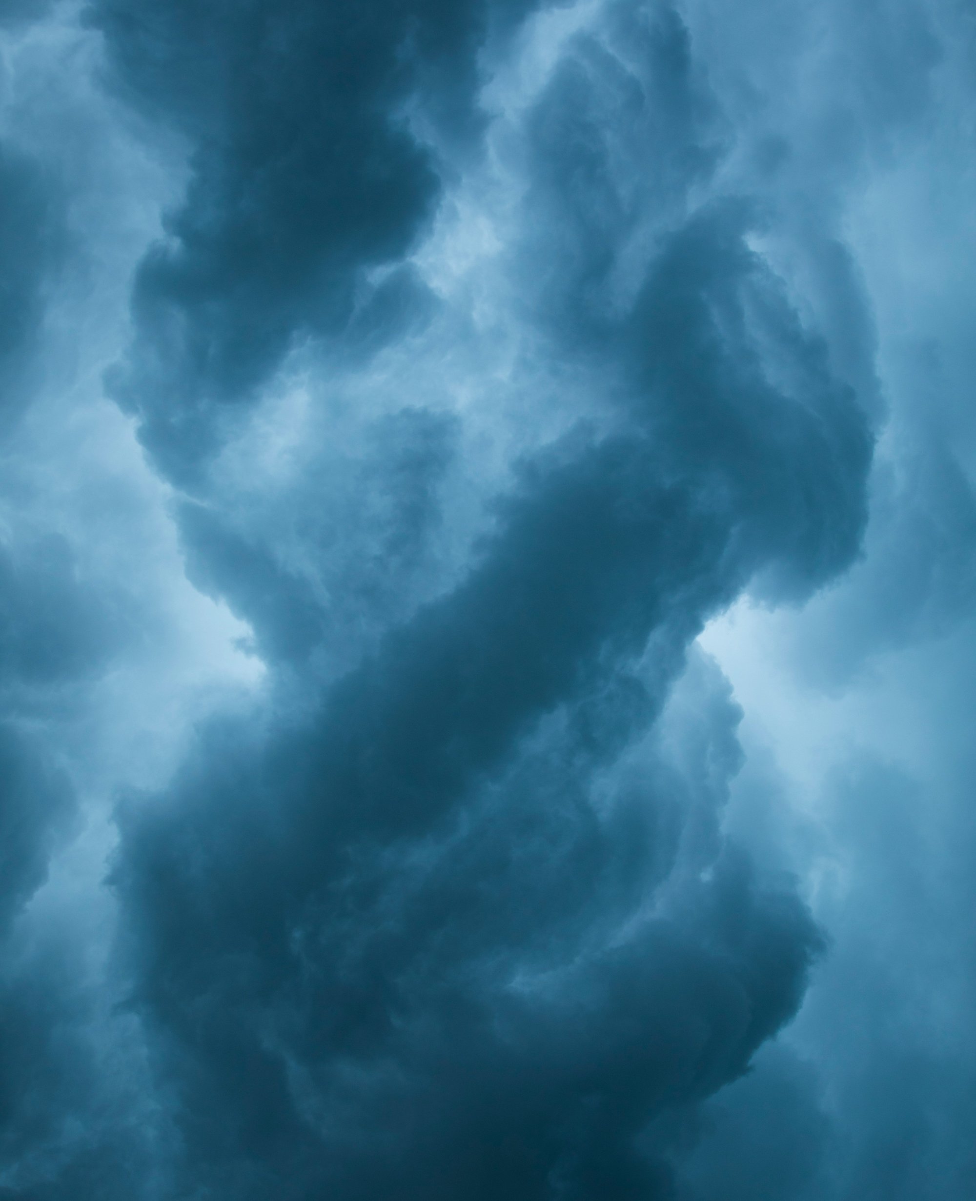 Dunkle, wirbelnde Gewitterwolken bedecken den Himmel und werden von einem schwachen Licht im Hintergrund beleuchtet, wodurch eine dramatische und stimmungsvolle Atmosphäre mit Blau- und Grautönen entsteht.