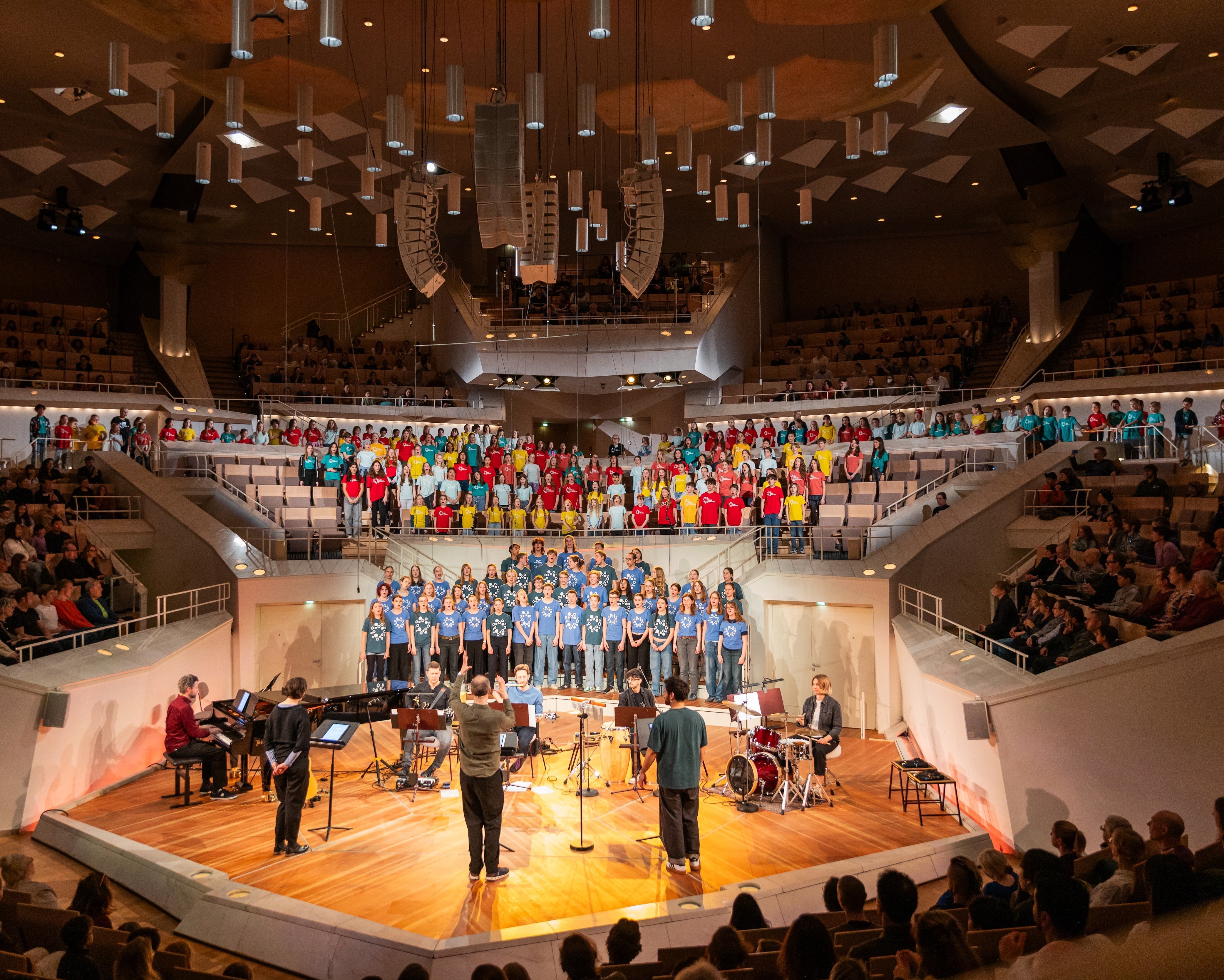 Ein Chor in farbenfrohen Kostümen tritt auf einer hell erleuchteten Bühne mit Musikern auf, während ein großes Publikum in einer modernen Konzerthalle mit Balkonen und geometrischen Deckenmustern zusieht.