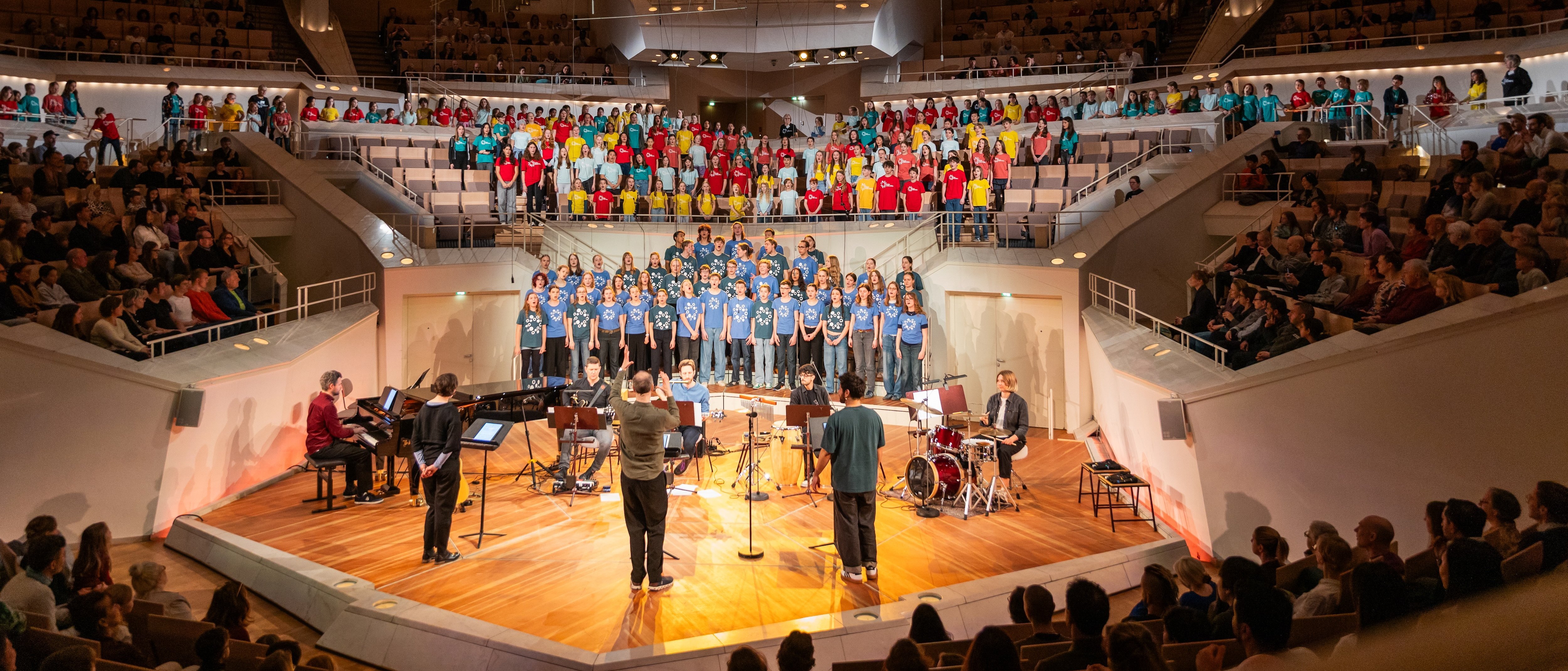 A large choir of people in colorful shirts sings on a brightly lit stage, accompanied by musicians, with an audience seated in a modern concert hall.