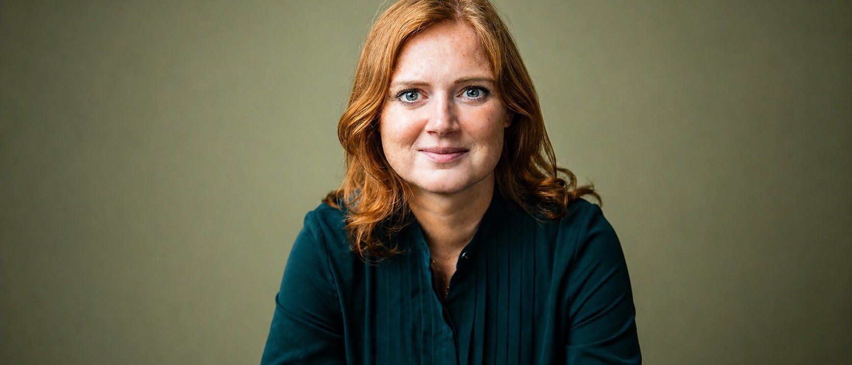 Eine Frau mit schulterlangen roten Haaren und einer dunkelgrünen Bluse sitzt vor einem schlichten, hellolivfarbenen Hintergrund und schaut mit einem sanften Lächeln direkt in die Kamera.
