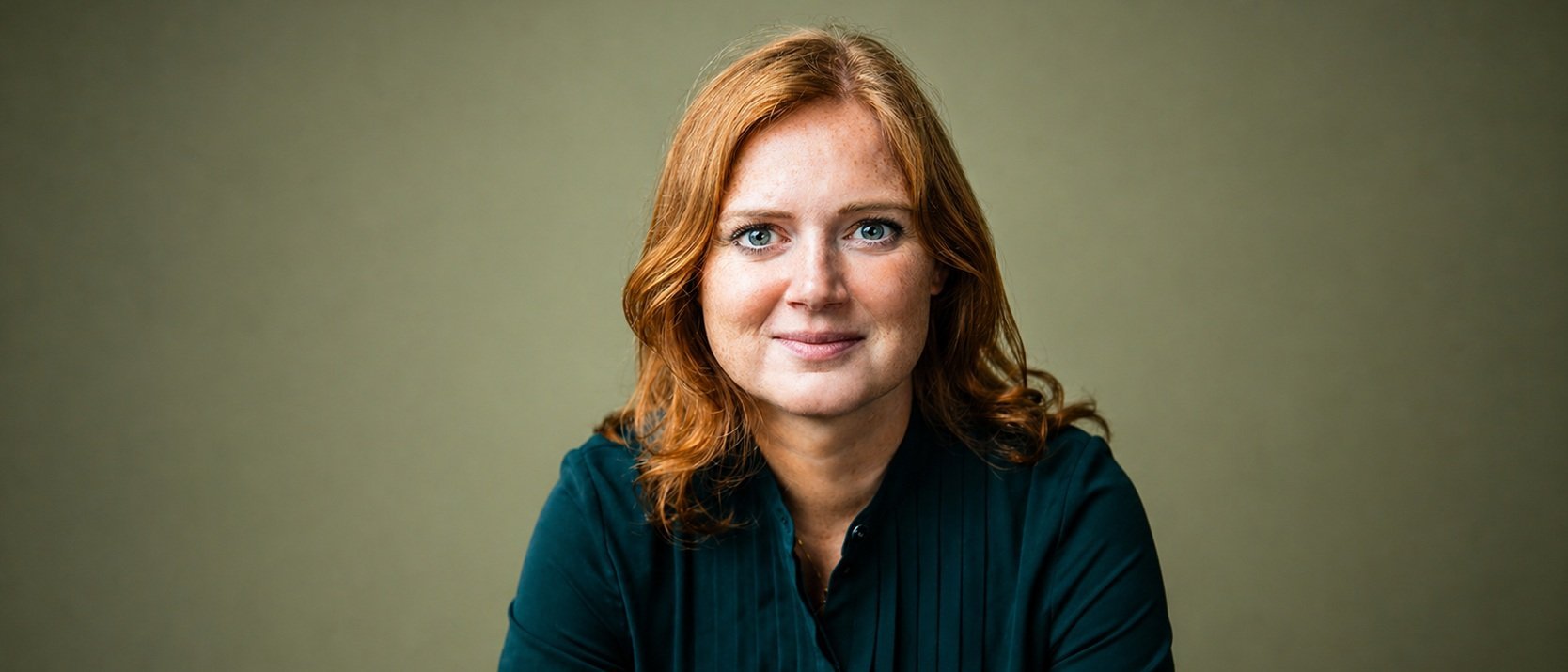 Eine Frau mit schulterlangen roten Haaren und einer dunkelgrünen Bluse sitzt vor einem schlichten, hellolivfarbenen Hintergrund und schaut mit einem sanften Lächeln direkt in die Kamera.