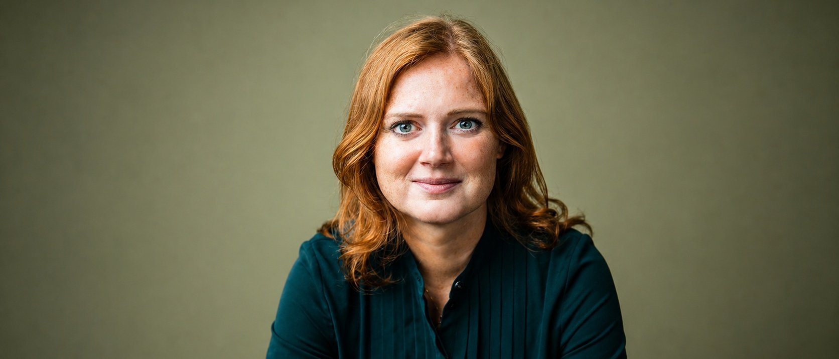 A woman with shoulder-length red hair wearing a dark green blouse sits against a plain, light olive background, looking directly at the camera with a neutral expression.