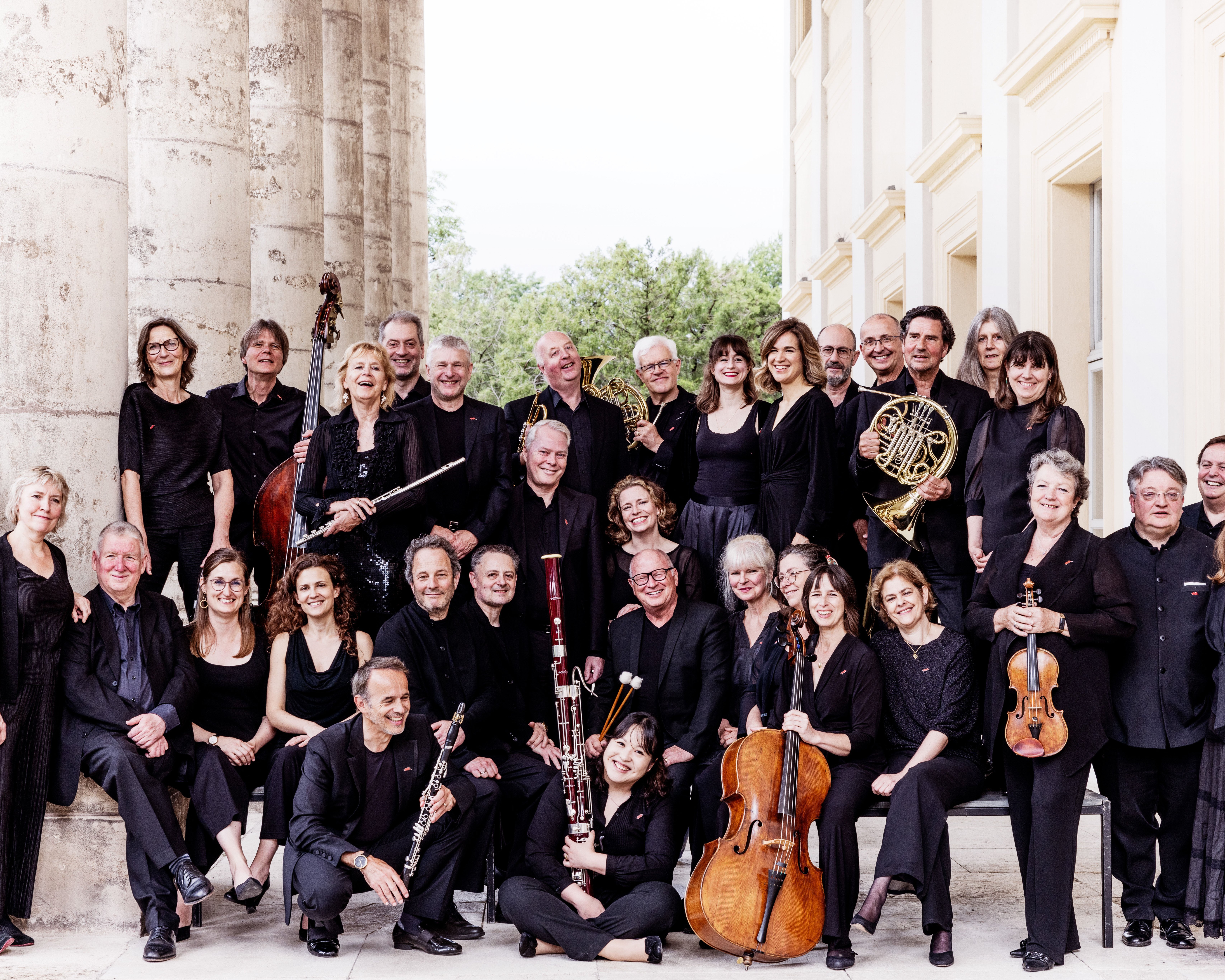 Das Chamber Orchestra of Europe posiert schwarz gekleidet vor einem eleganten Gebäude. Sie stehen auf Steinstufen und halten verschiedene Instrumente in den Händen, darunter Geigen, Klarinetten, ein Horn und ein Fagott.