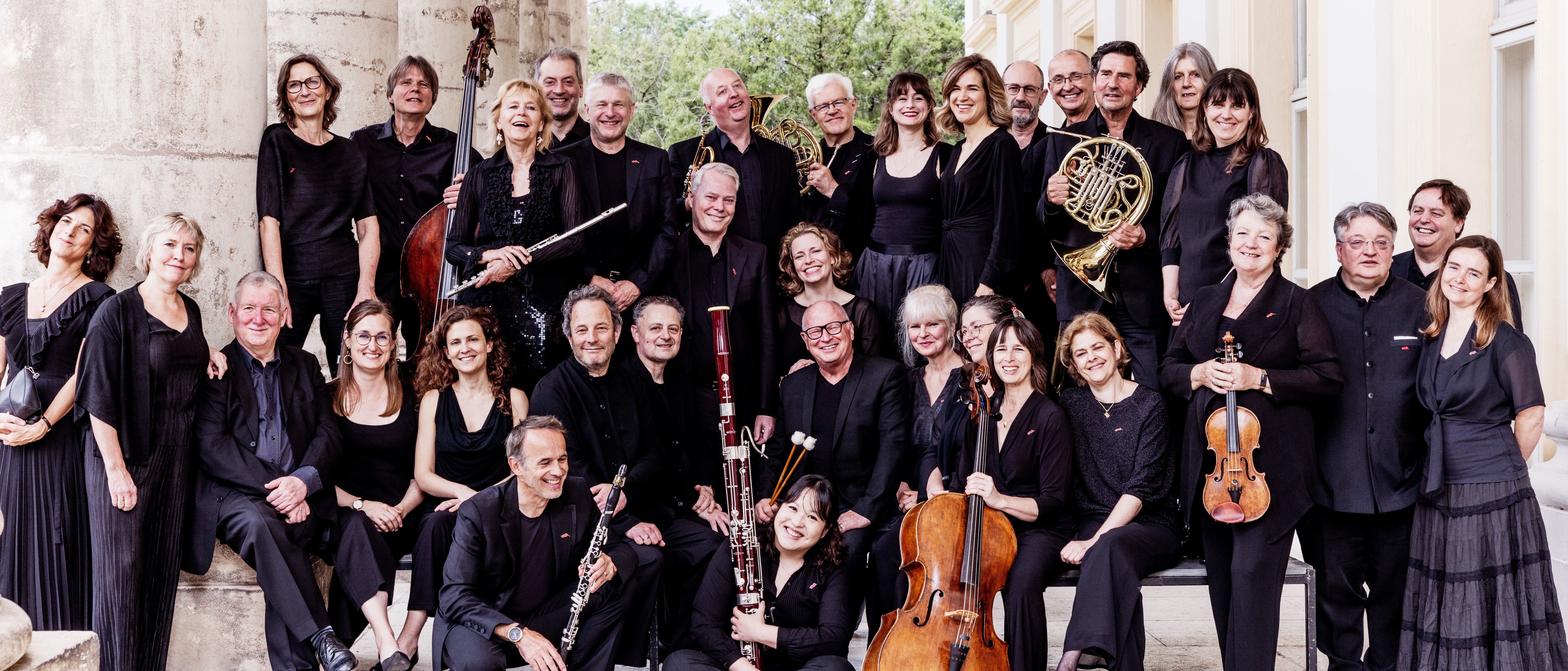 Das Chamber Orchestra of Europe posiert schwarz gekleidet vor einem eleganten Gebäude. Sie stehen auf Steinstufen und halten verschiedene Instrumente in den Händen, darunter Geigen, Klarinetten, ein Horn und ein Fagott.