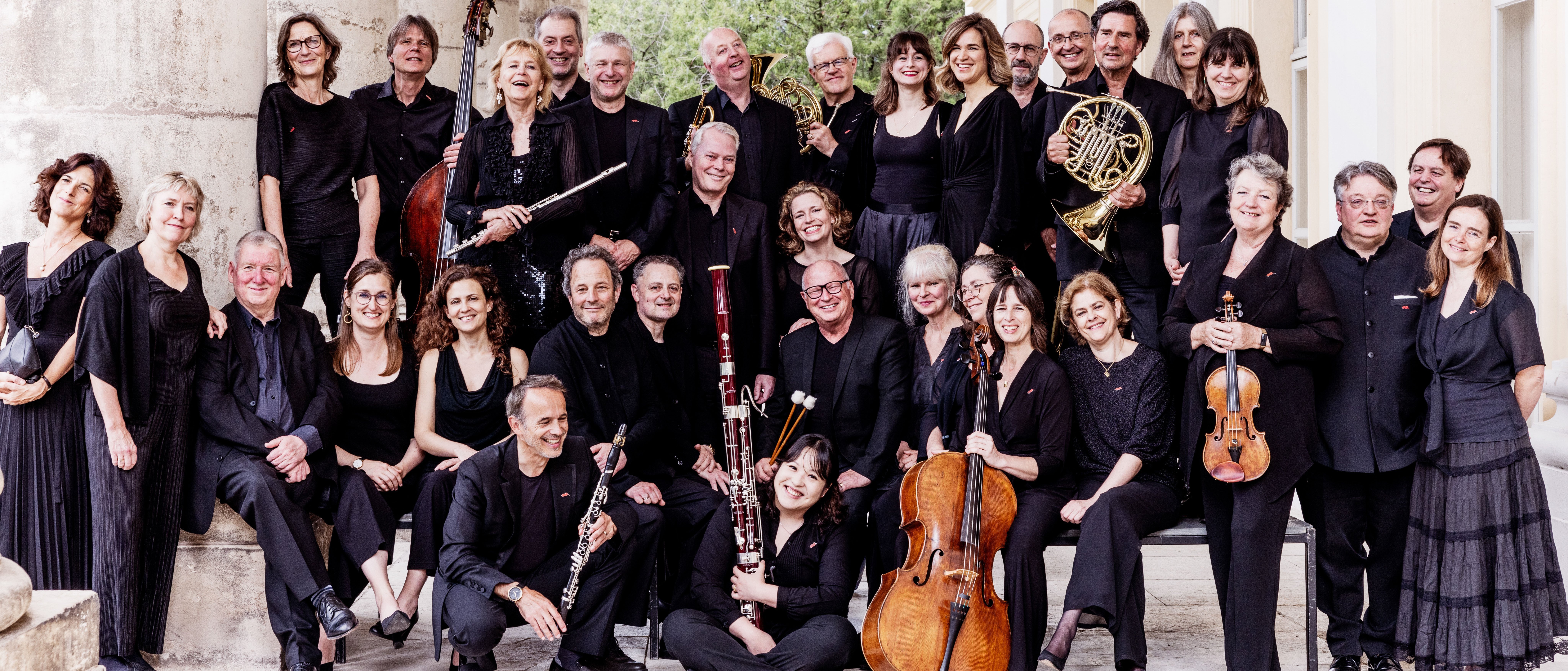 Das Chamber Orchestra of Europe posiert schwarz gekleidet vor einem eleganten Gebäude. Sie stehen auf Steinstufen und halten verschiedene Instrumente in den Händen, darunter Geigen, Klarinetten, ein Horn und ein Fagott.