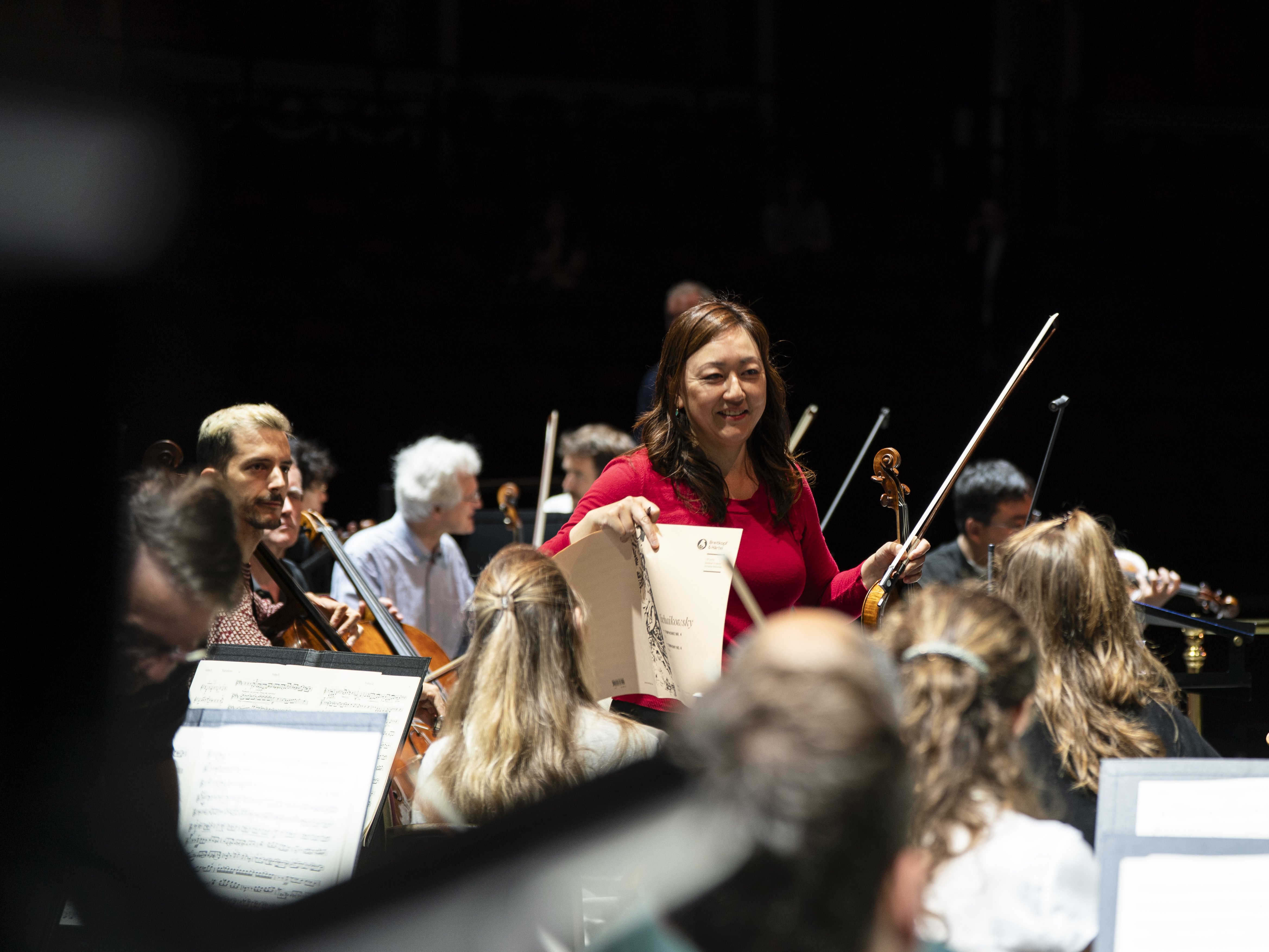 Eine Frau steht lächelnd mit einer Geige und Notenblättern in der Hand inmitten eines sitzenden Orchesters, umgeben von Musikern, die auf einer schwach beleuchteten Bühne verschiedene Instrumente spielen.